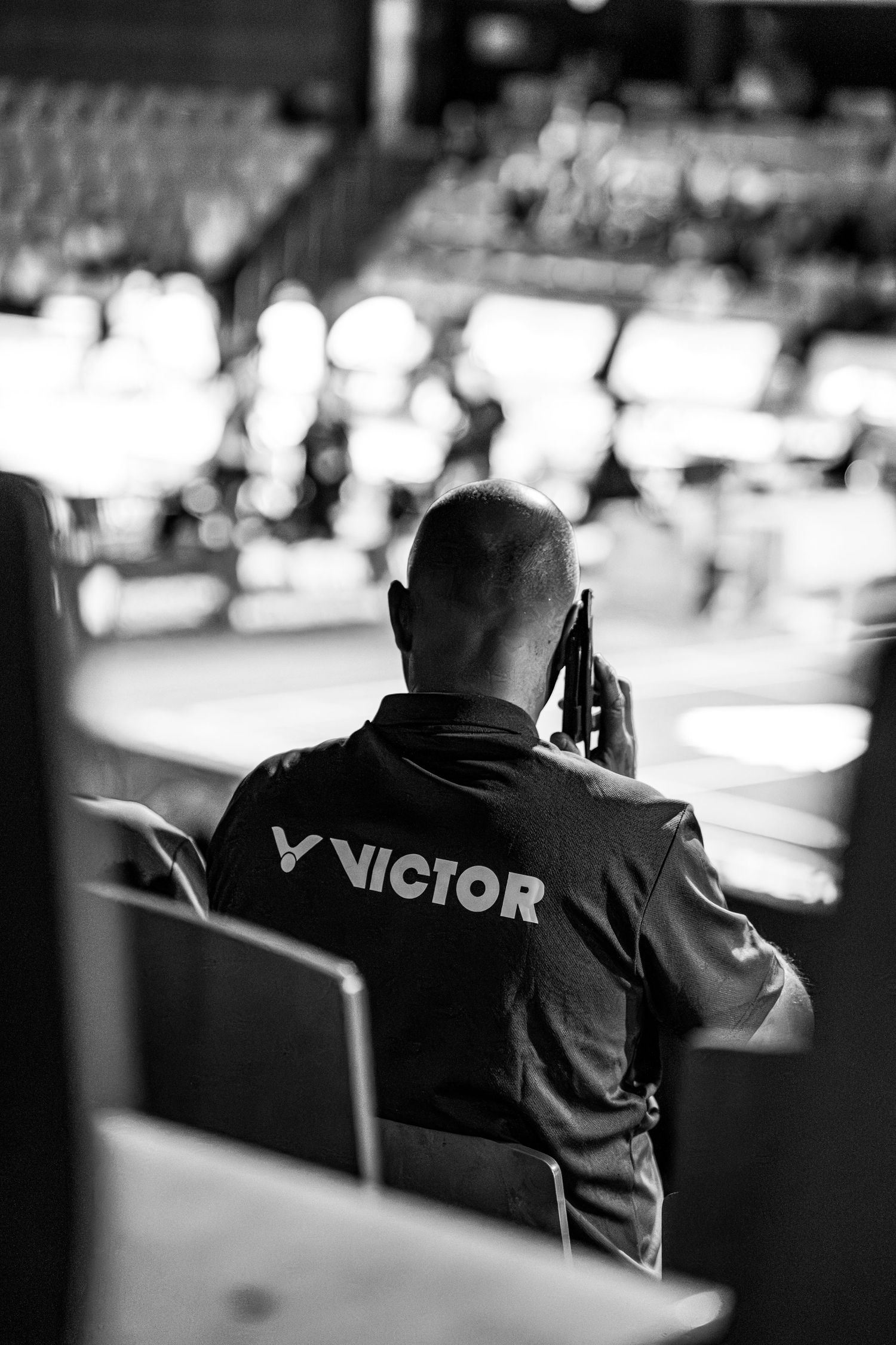 Man wearing Victor sports jacket talks on phone at a badminton tournament in black and white.