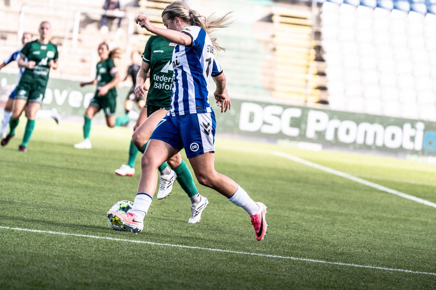 Two female soccer players compete for the ball during a match between blue-white and green-kitted teams.