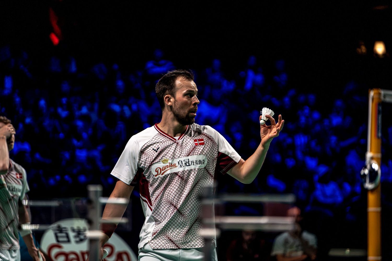 Professional badminton player holding shuttlecock preparing to serve during competitive indoor tournament match.