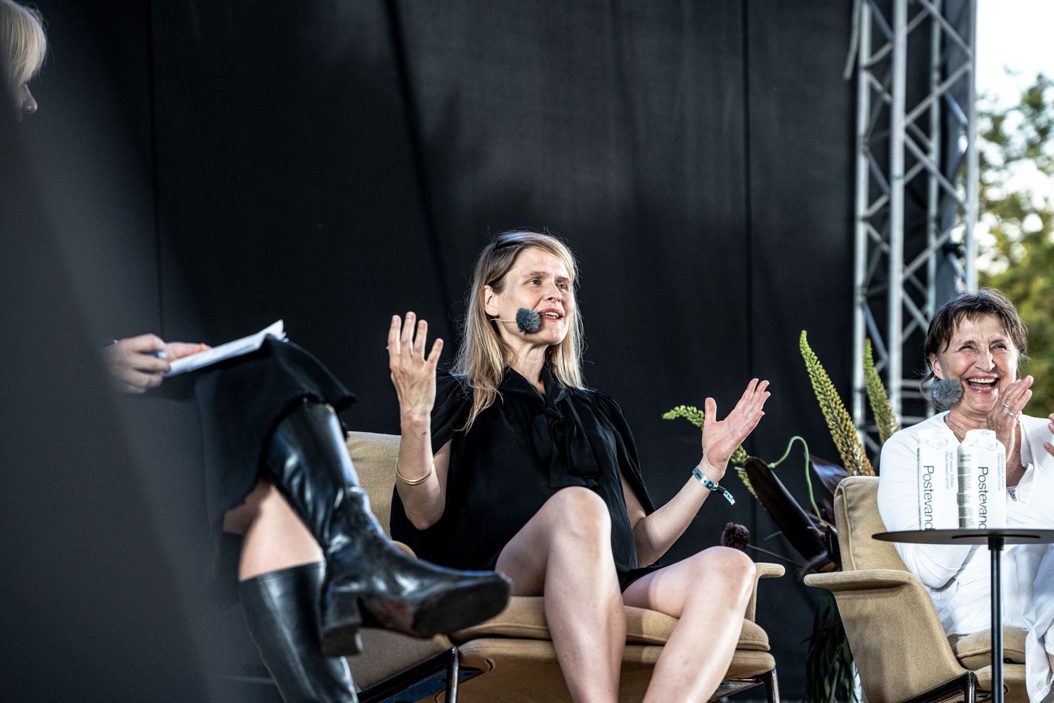 Woman in black dress gesturing with hands while speaking at an outdoor stage panel discussion.