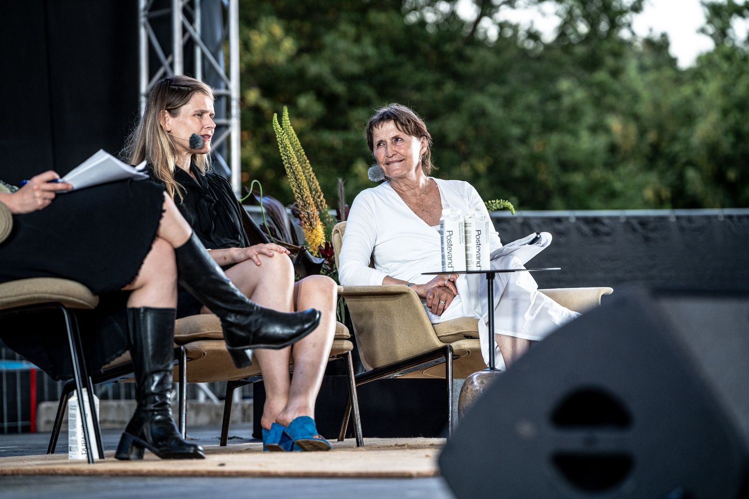 Two women sitting on an outdoor stage during a panel discussion, surrounded by greenery.