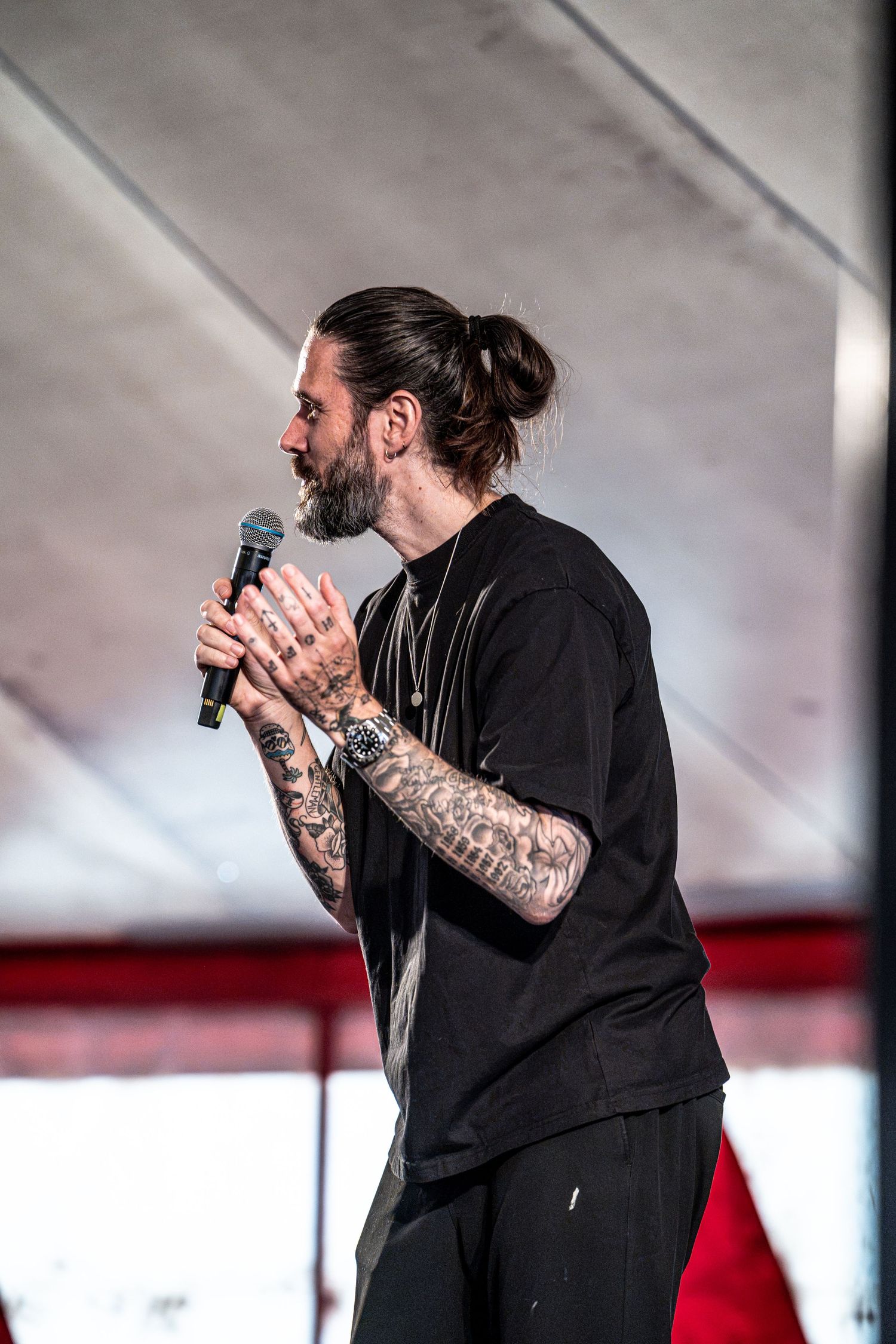 Tattooed bearded male singer holds microphone and gestures on stage at outdoor music festival.