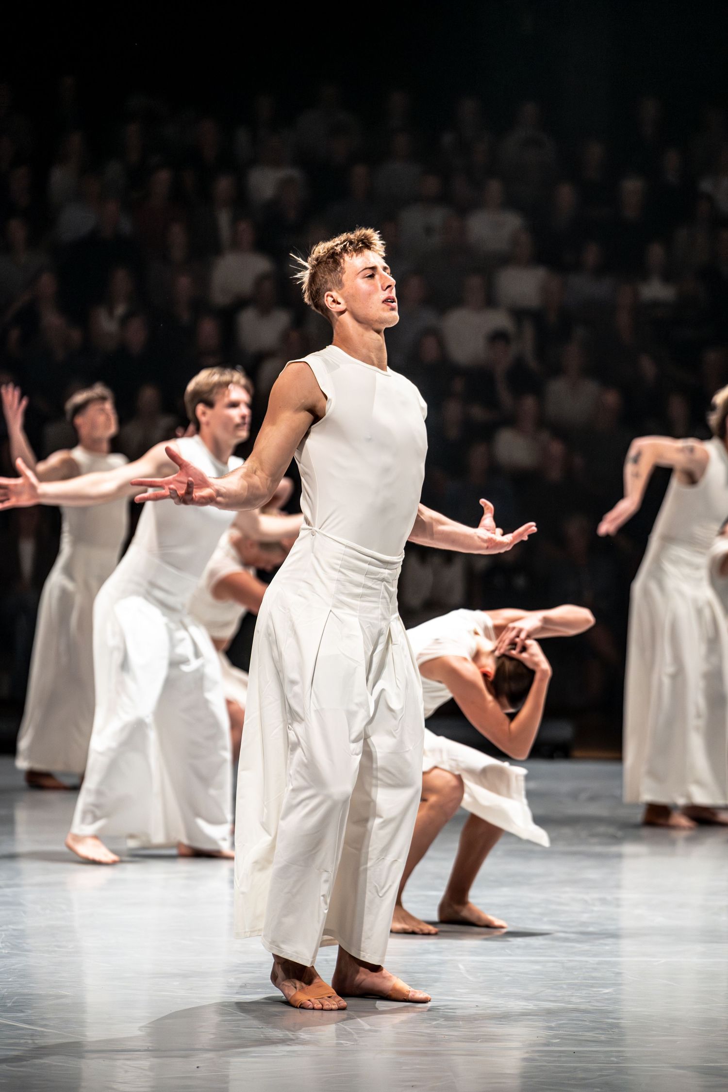 Male contemporary dancer in white outfit performs on stage with ensemble dancers behind him.