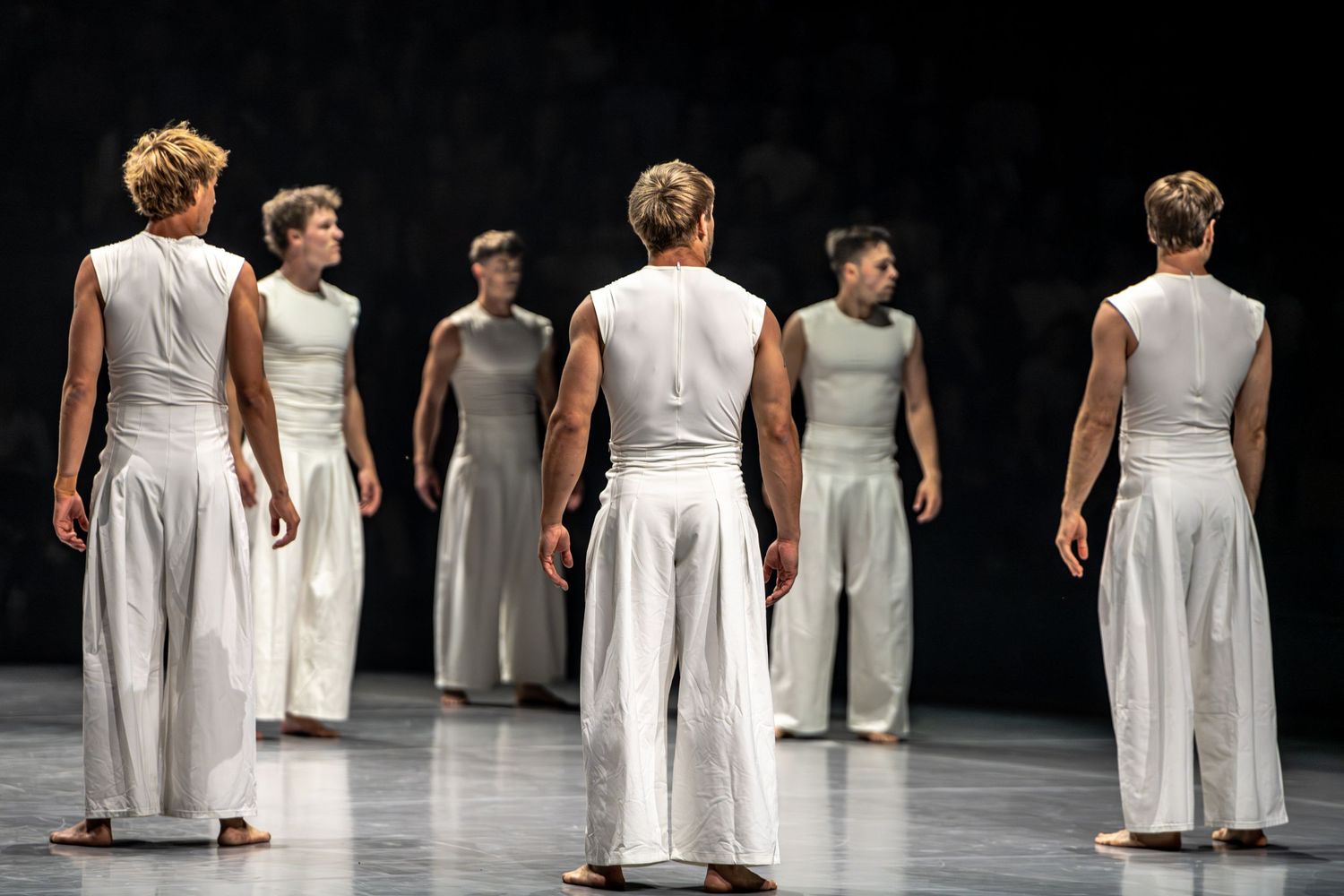 Group of contemporary dancers in white costumes stand on dark stage during performance.