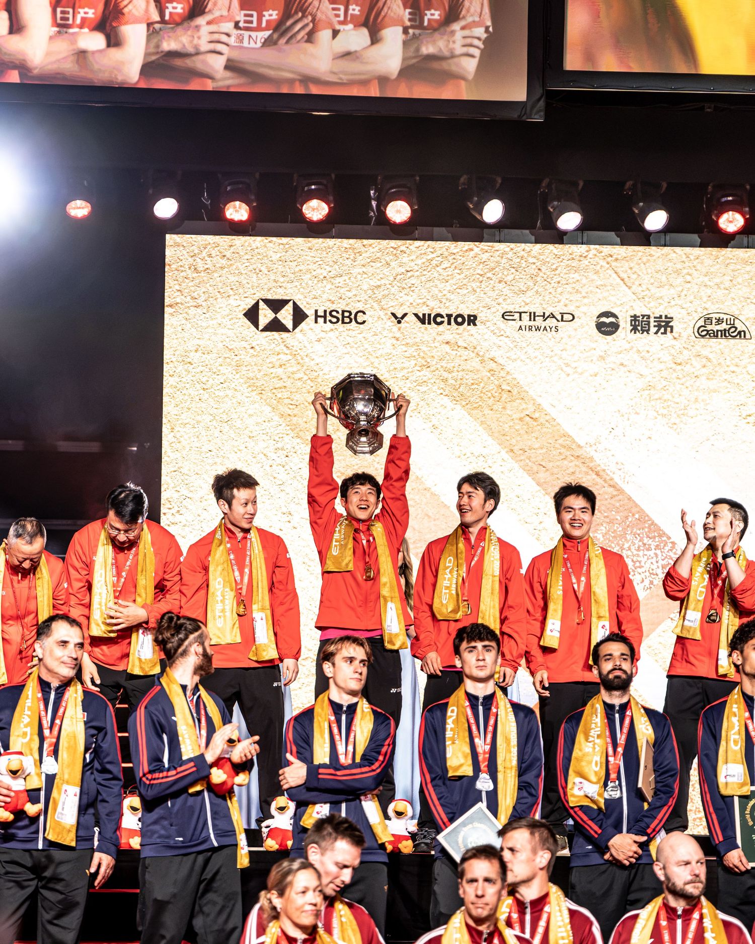 Team China captain lifts trophy on stage as teammates celebrate badminton championship win.