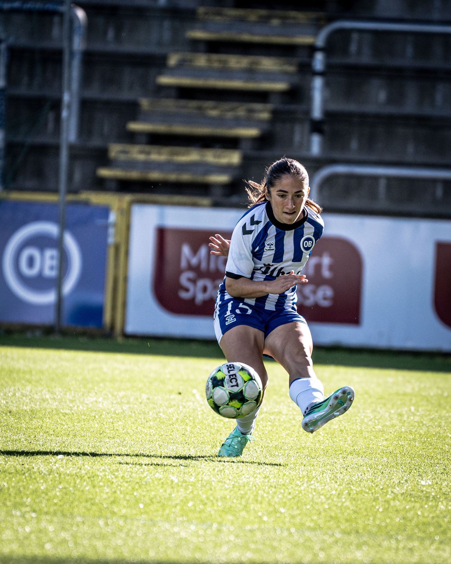 Female soccer player in blue-white Hummel kit strikes ball during women's football match.