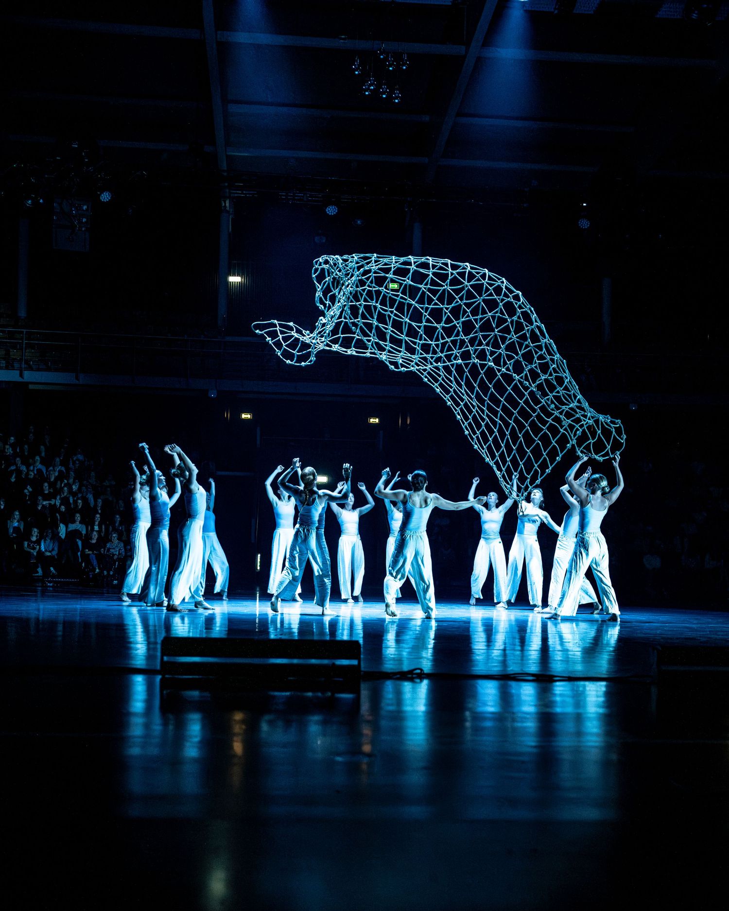 Dancers in white carry a glowing wire-frame elephant sculpture under blue stage lighting.