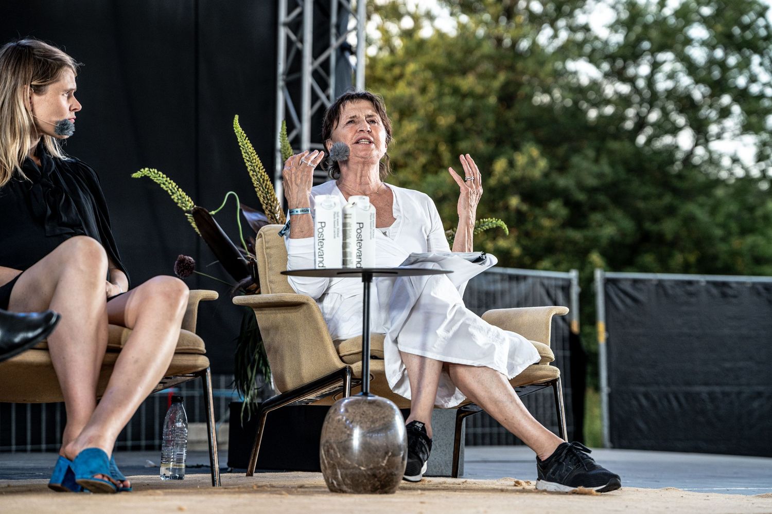 Woman in white dress speaking animatedly at an outdoor panel event on a wooden stage.