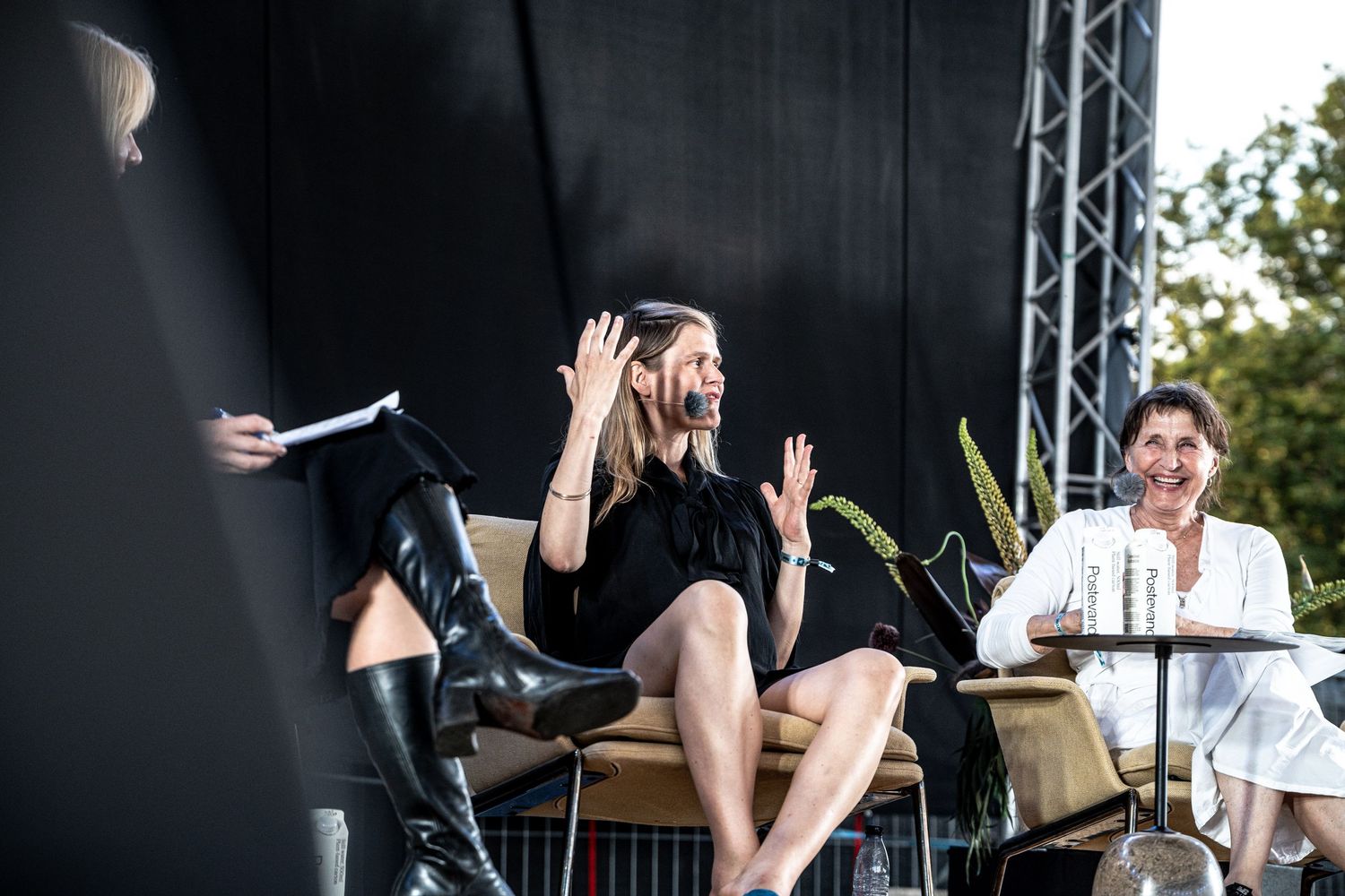 Woman in black dress raising both hands expressively during an outdoor stage panel discussion.
