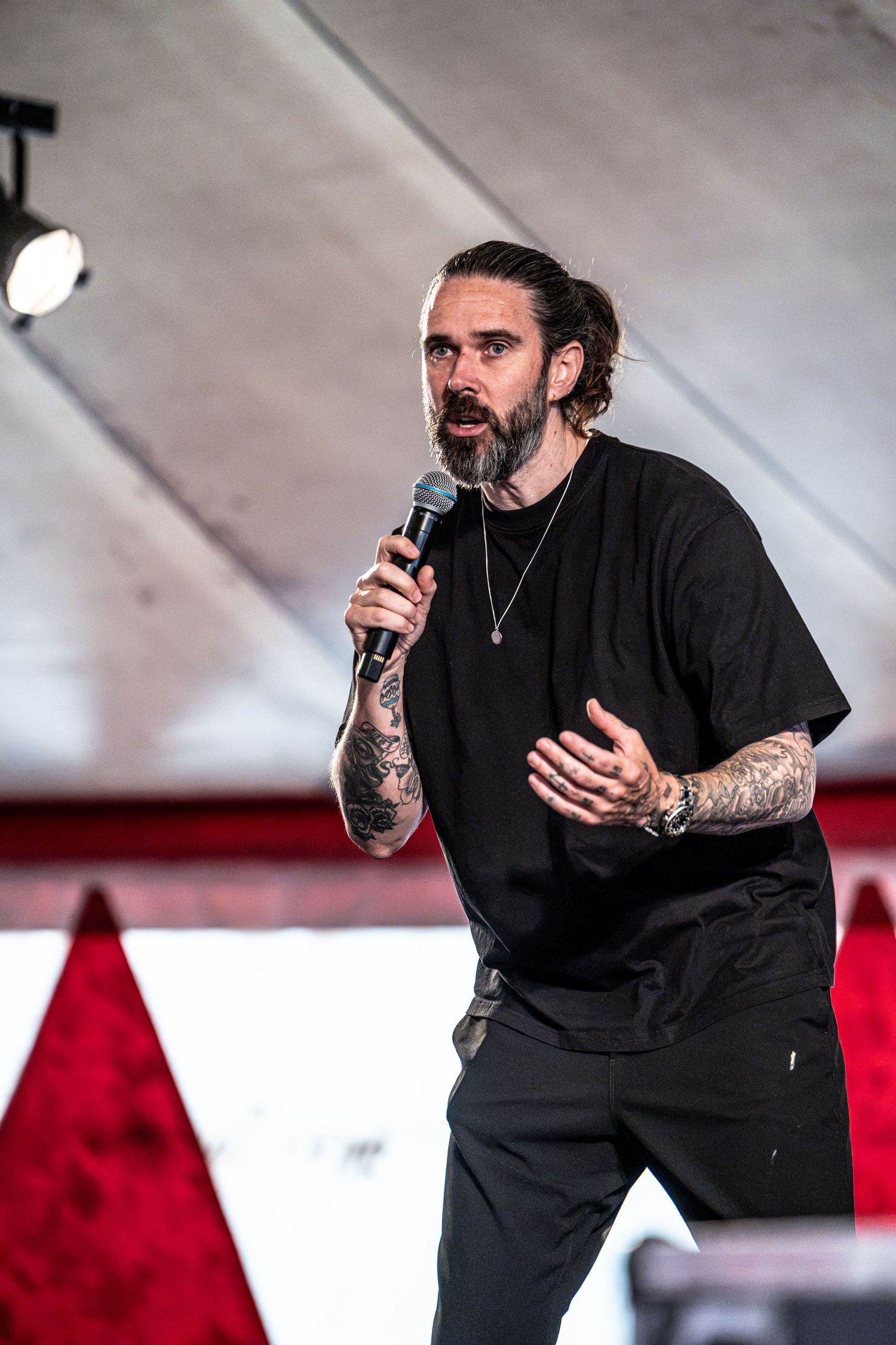 Tattooed male performer in black outfit speaking into microphone on outdoor stage.