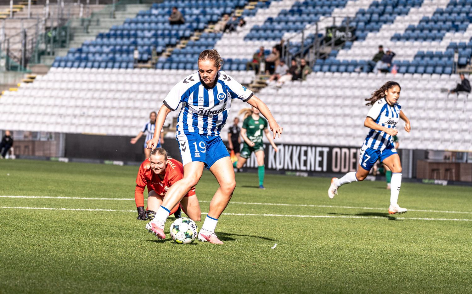 Women's footballer number 19 steps over sliding goalkeeper while maintaining ball control during match.