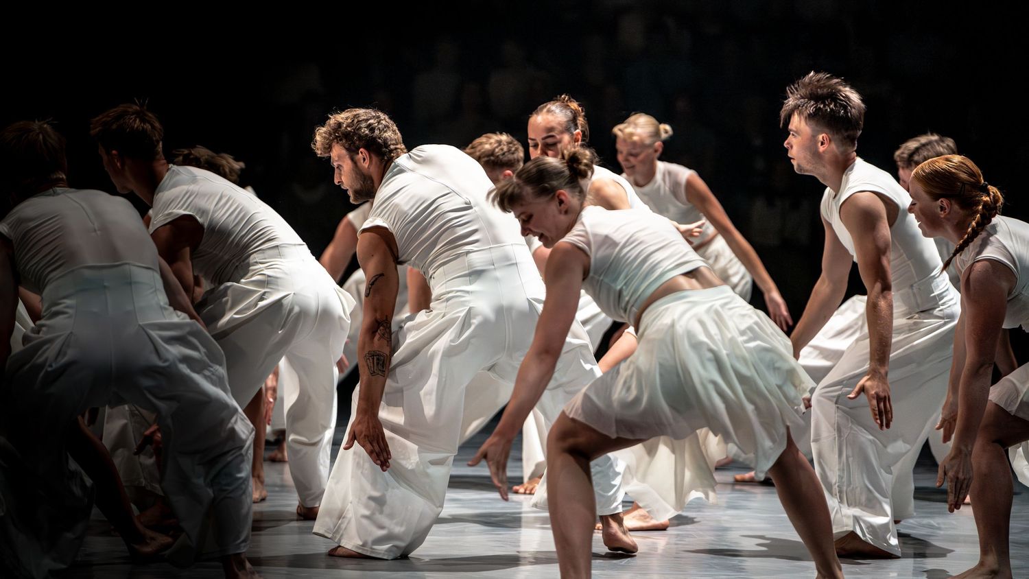 Contemporary dancers in white costumes perform an expressive group routine on a dimly lit stage.