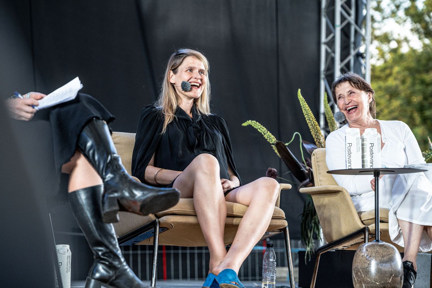 Two women laughing on stage at an outdoor panel event, one wearing black dress with microphone headset.