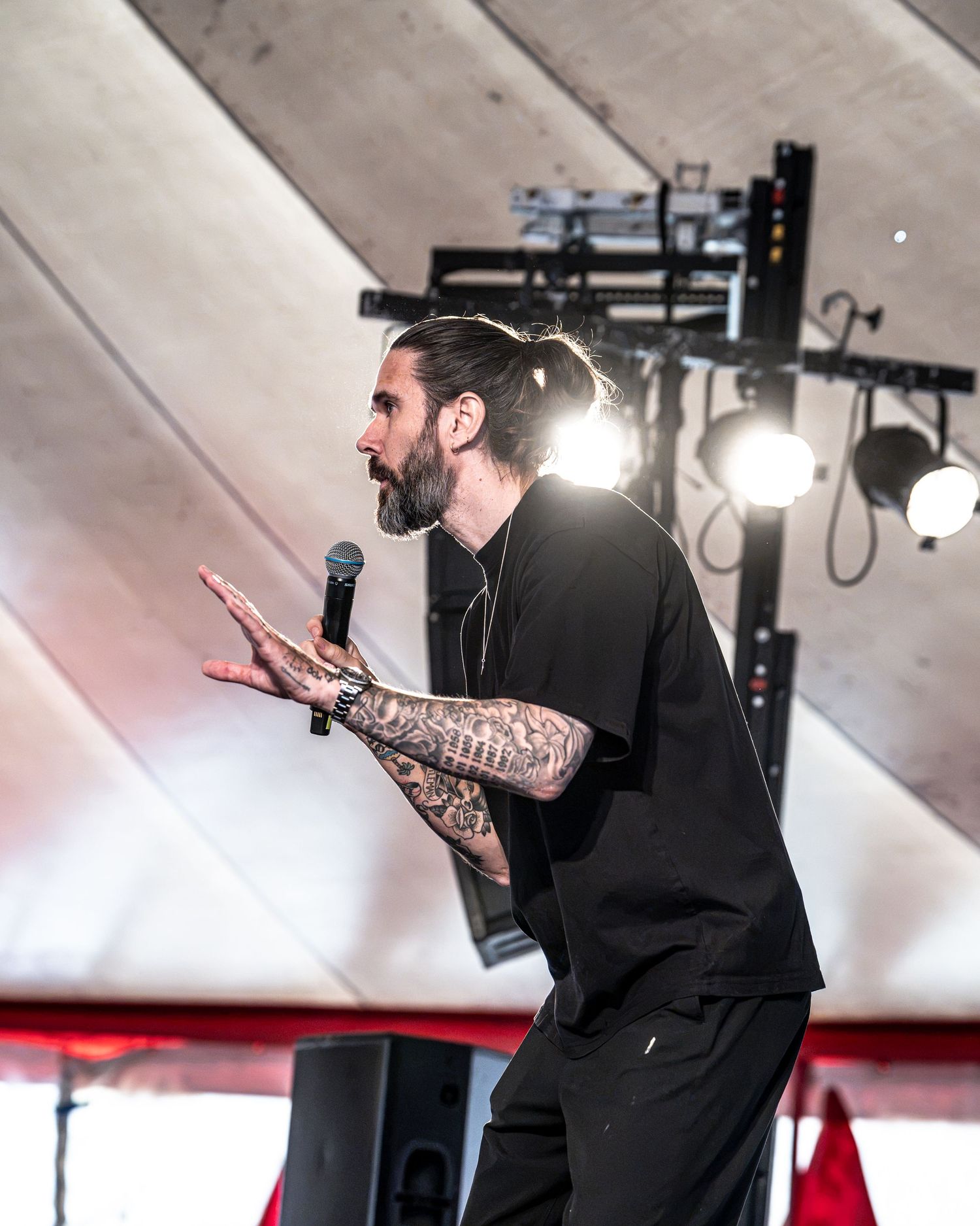 Bearded tattooed male performer gestures while speaking into microphone on stage at music festival.