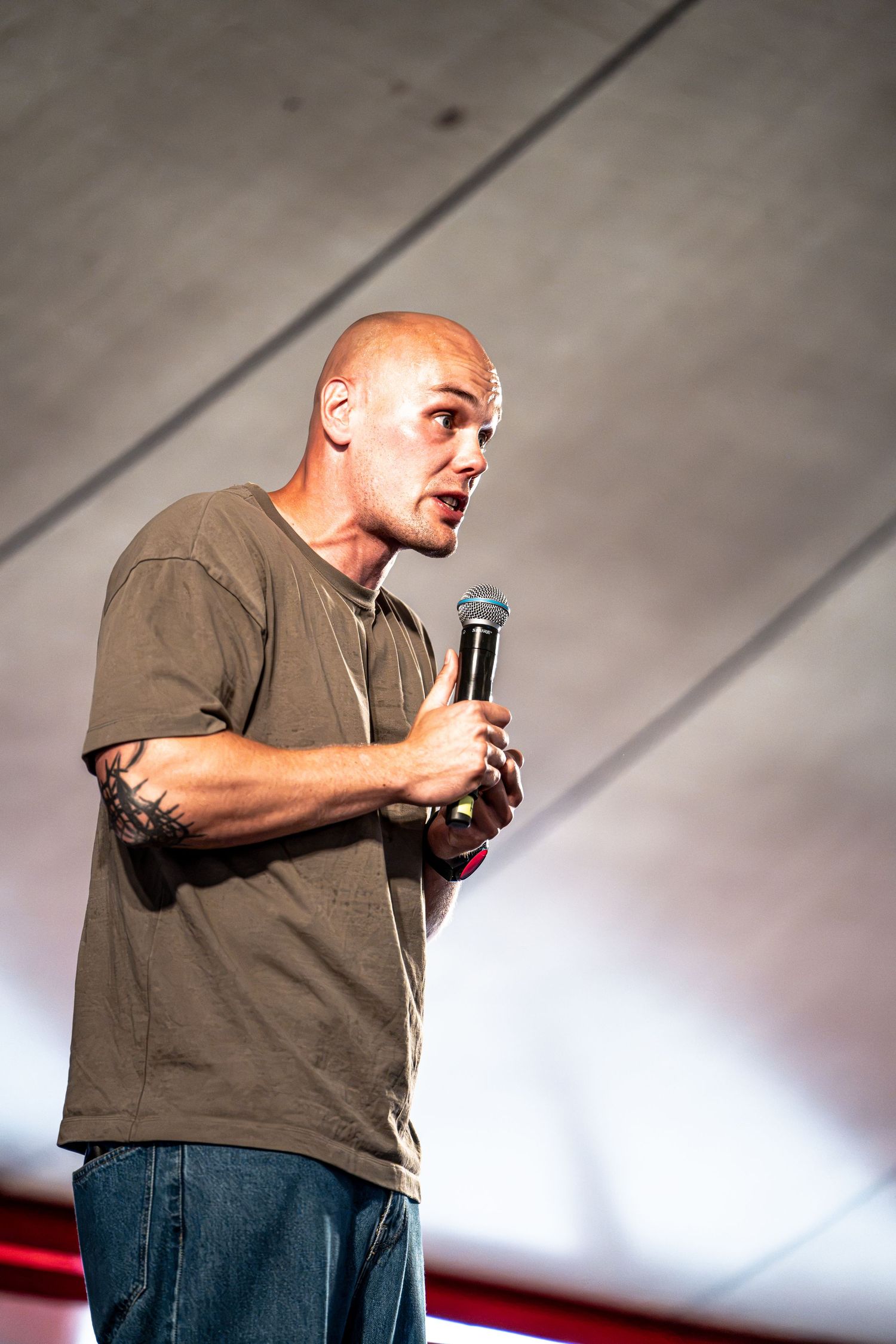 Bald tattooed man speaking intensely into a microphone on an outdoor stage.