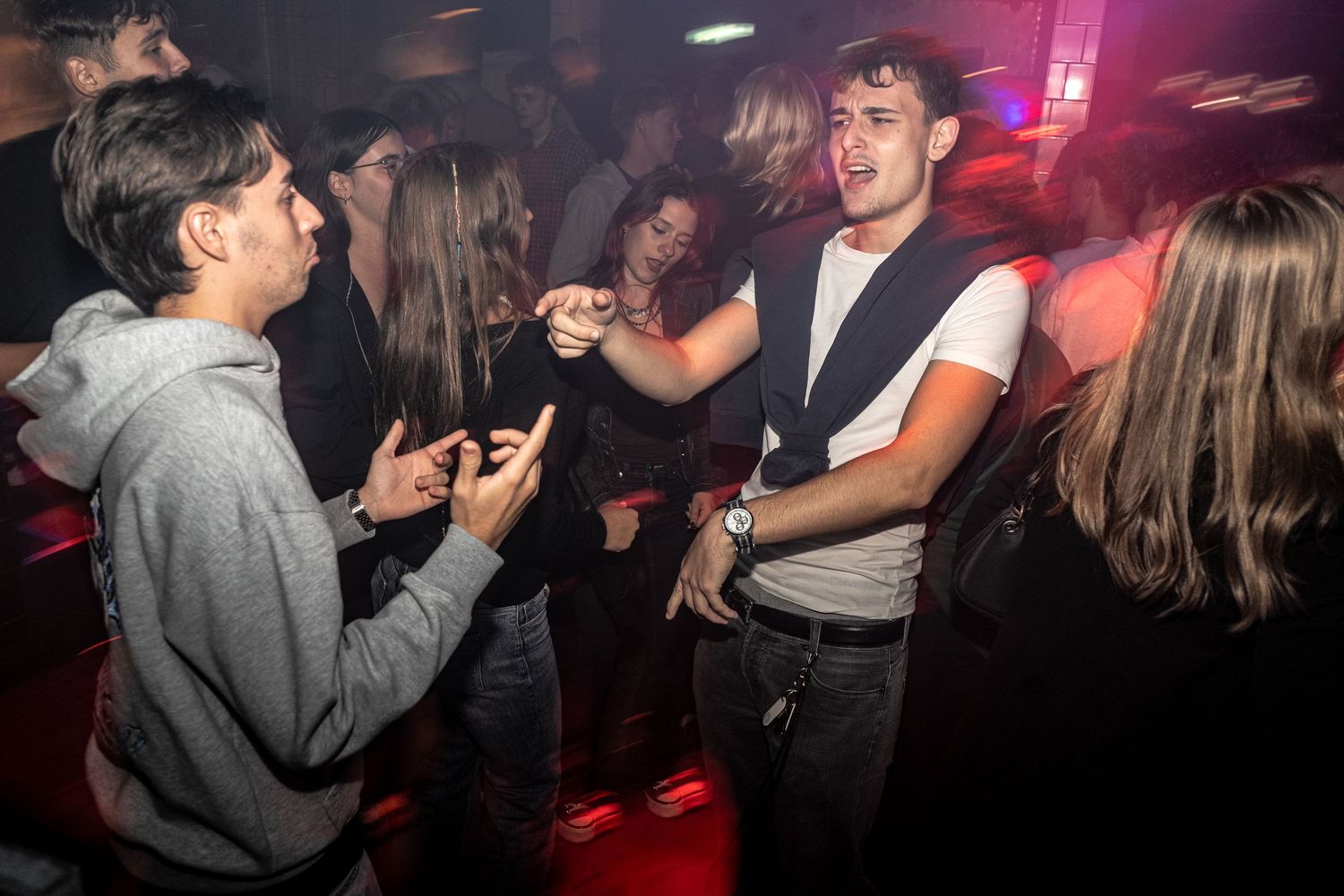 Young men dancing and gesturing in a packed nightclub under moody red and purple lighting, surrounded by a lively crowd.