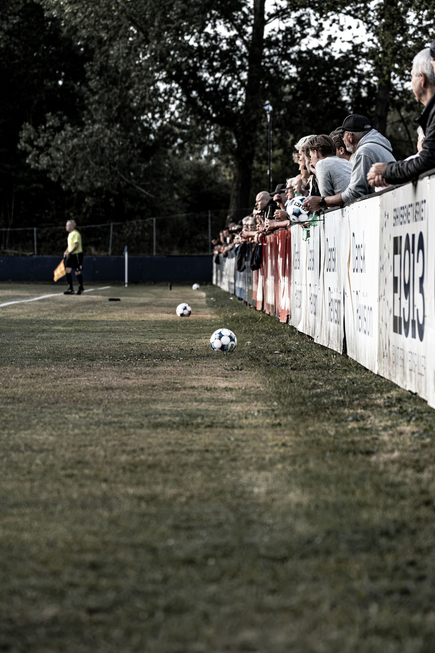 Soccer balls line the sideline as spectators watch a match from behind advertising boards.