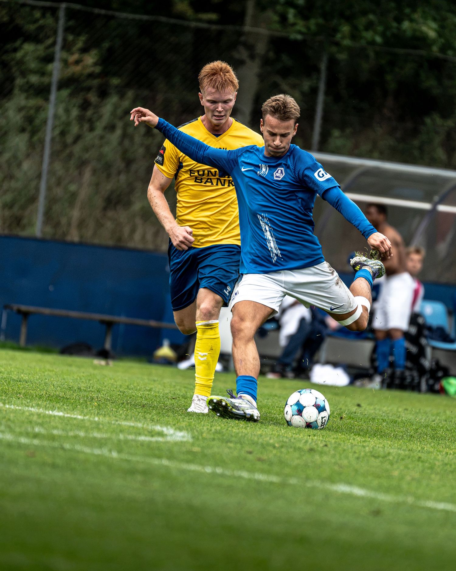 Soccer player in blue uniform kicking the ball while defended by yellow-jersey opponent.