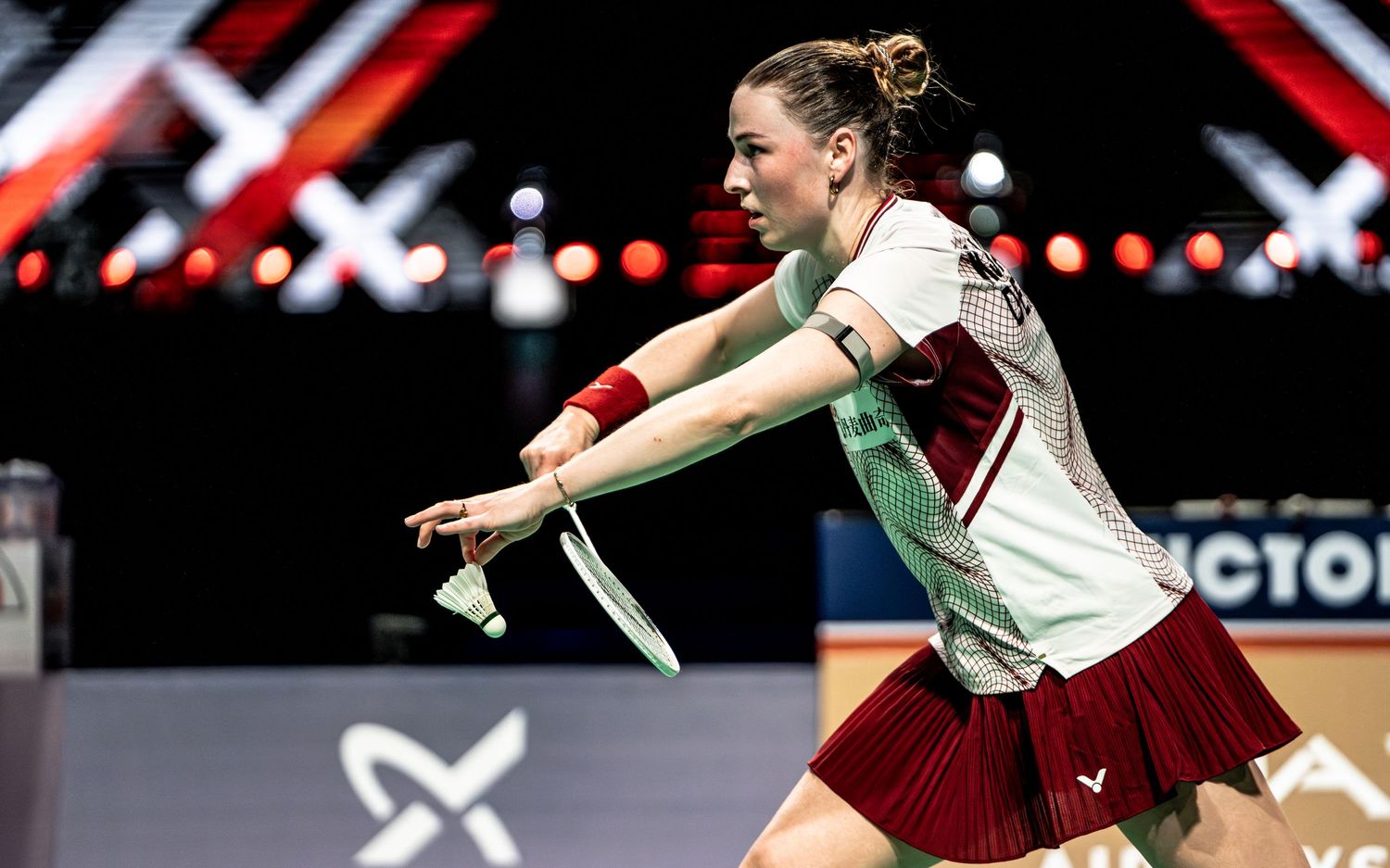 Female badminton player in red and white jersey reaching forward to return shuttlecock during tournament.