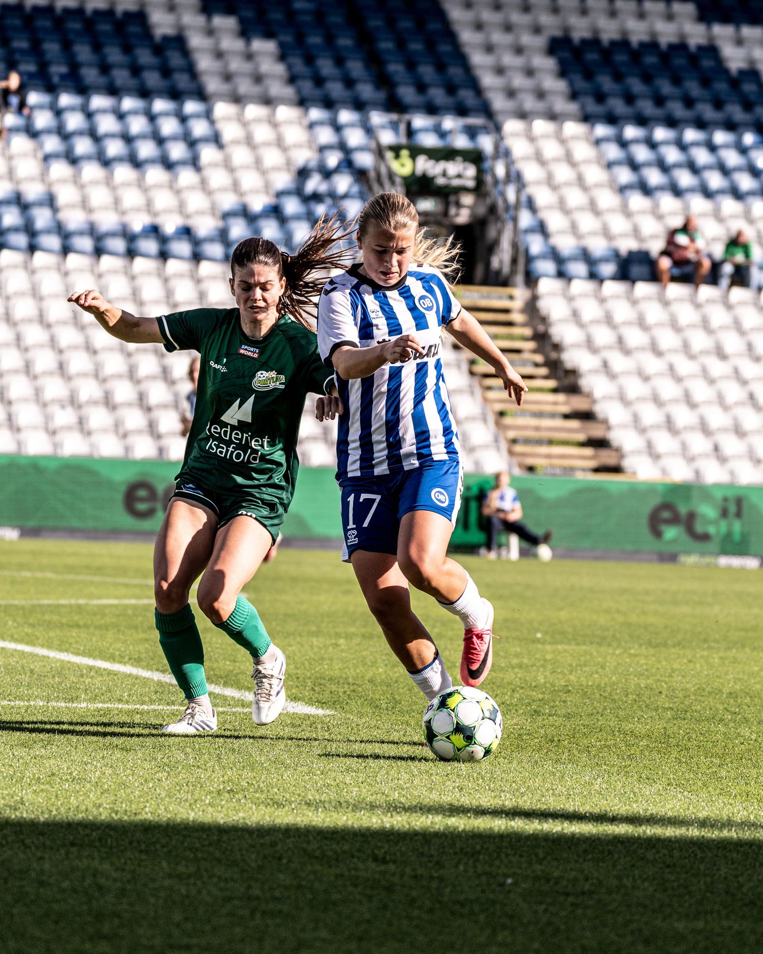 Player #17 in blue and white battles for the ball against a green-kitted opponent in a women's soccer match.