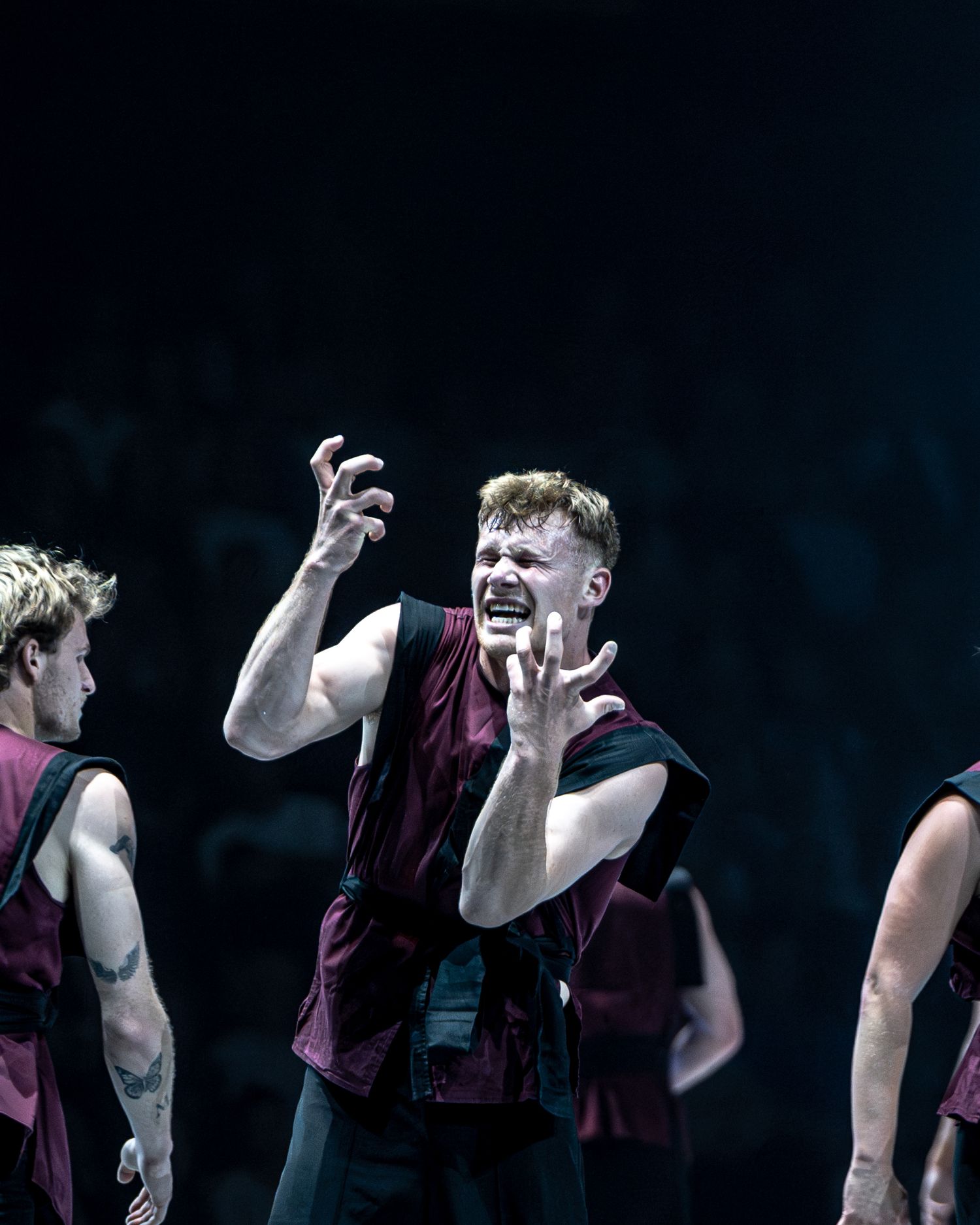 Expressive male dancer in burgundy vest performing passionately with ensemble on dark stage.