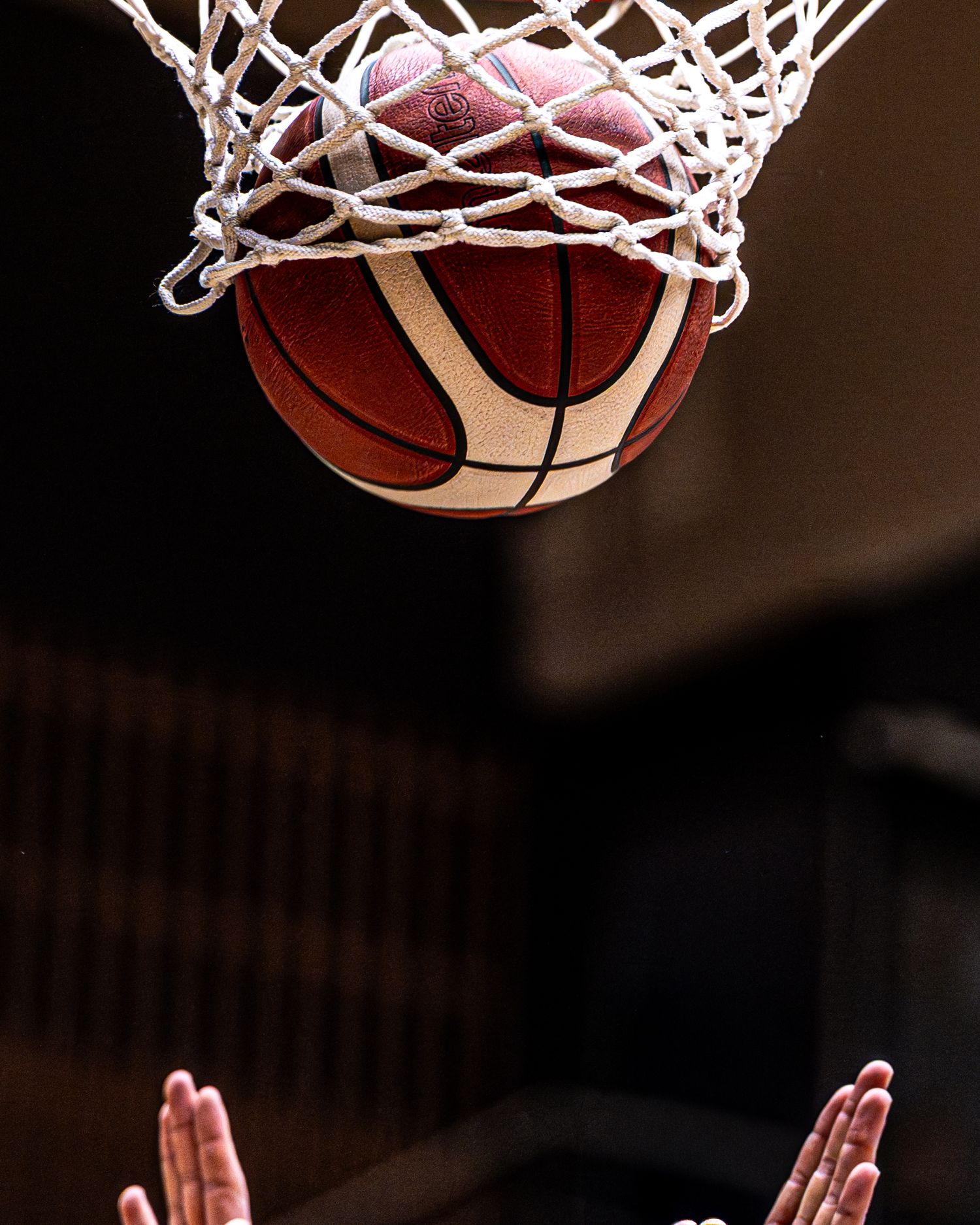 Basketball passing through net hoop with hands reaching up from below.