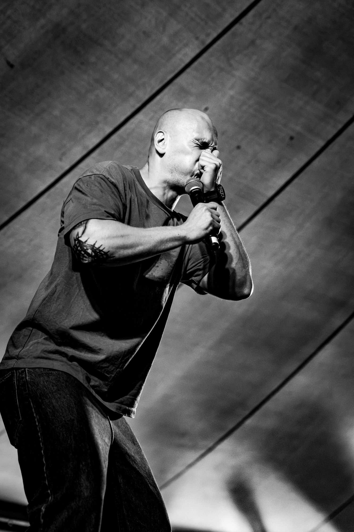 Black and white photo of a bald male singer leaning forward intensely into a microphone on stage.