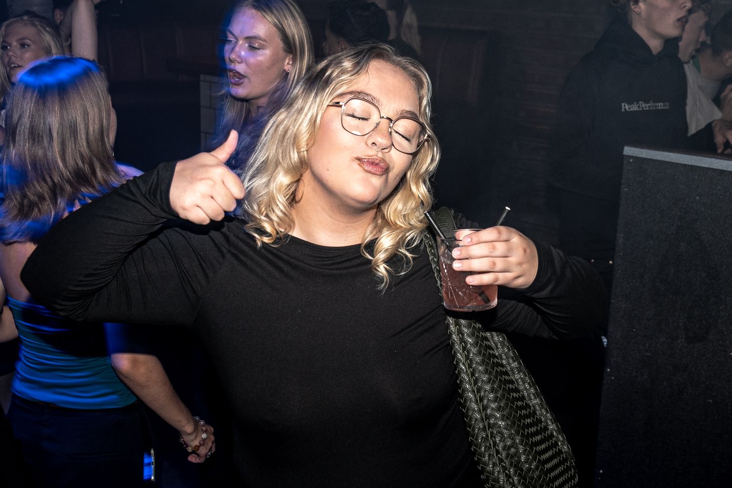 Woman with wavy blonde hair and round glasses gives a thumbs up while holding a drink at a lively nightclub party.