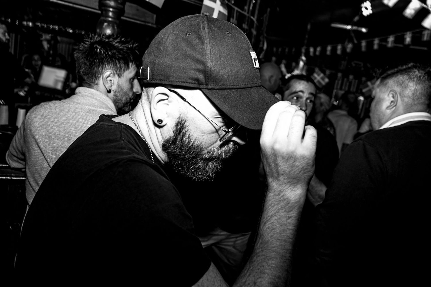 Black and white photo of a bearded man in a baseball cap and glasses raising his fist while dancing in a crowded, dimly lit bar.