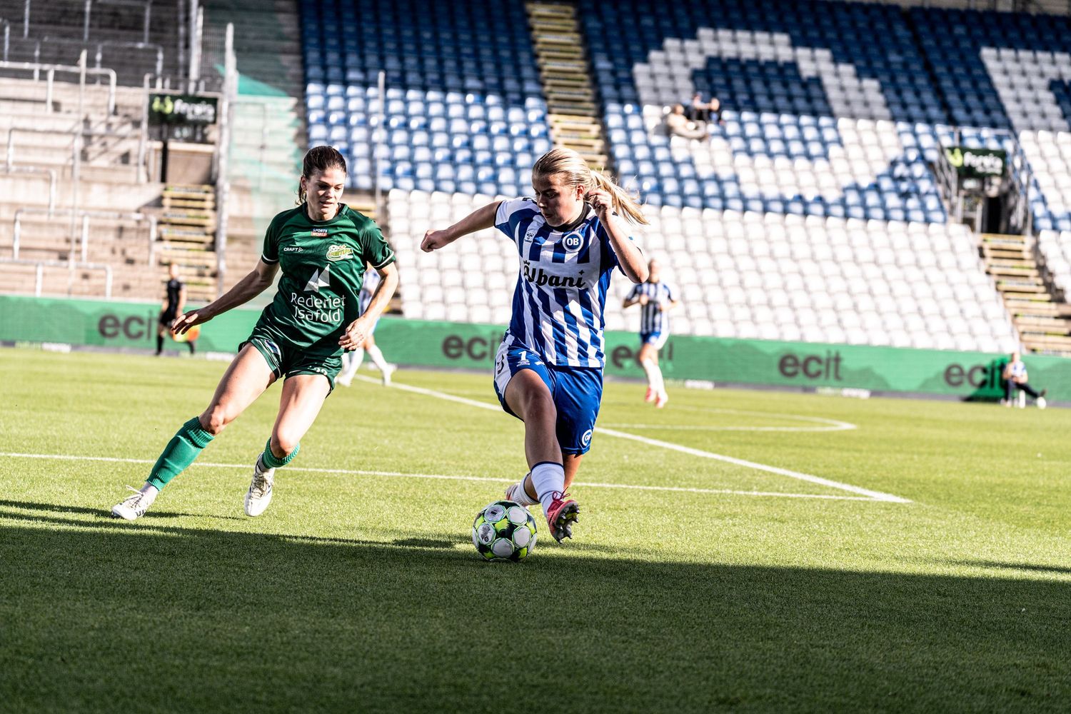 Female footballer in blue and white kit sprints past green jersey opponent during stadium match.