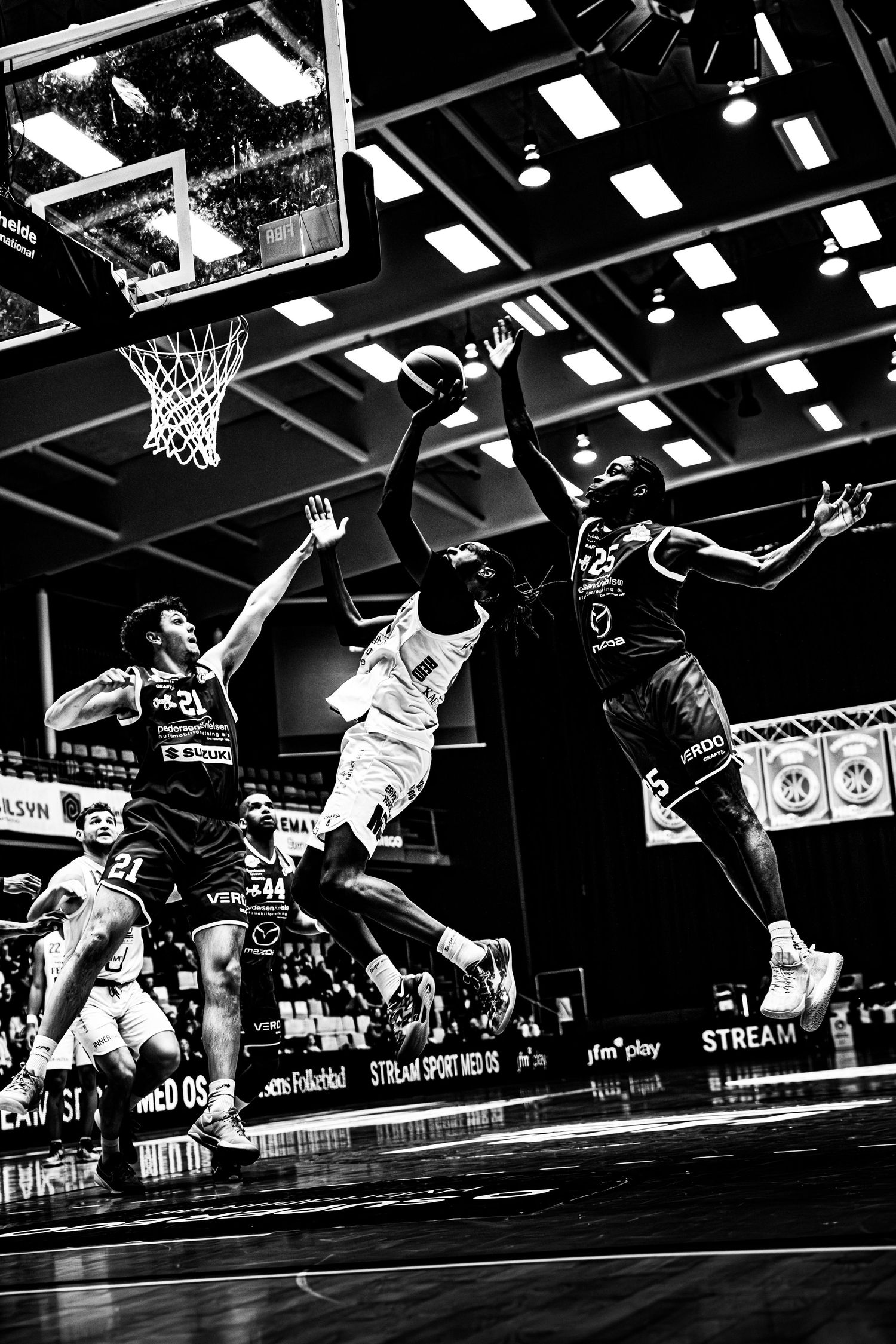 Black and white action shot of basketball players competing for the ball near the hoop in an indoor arena.