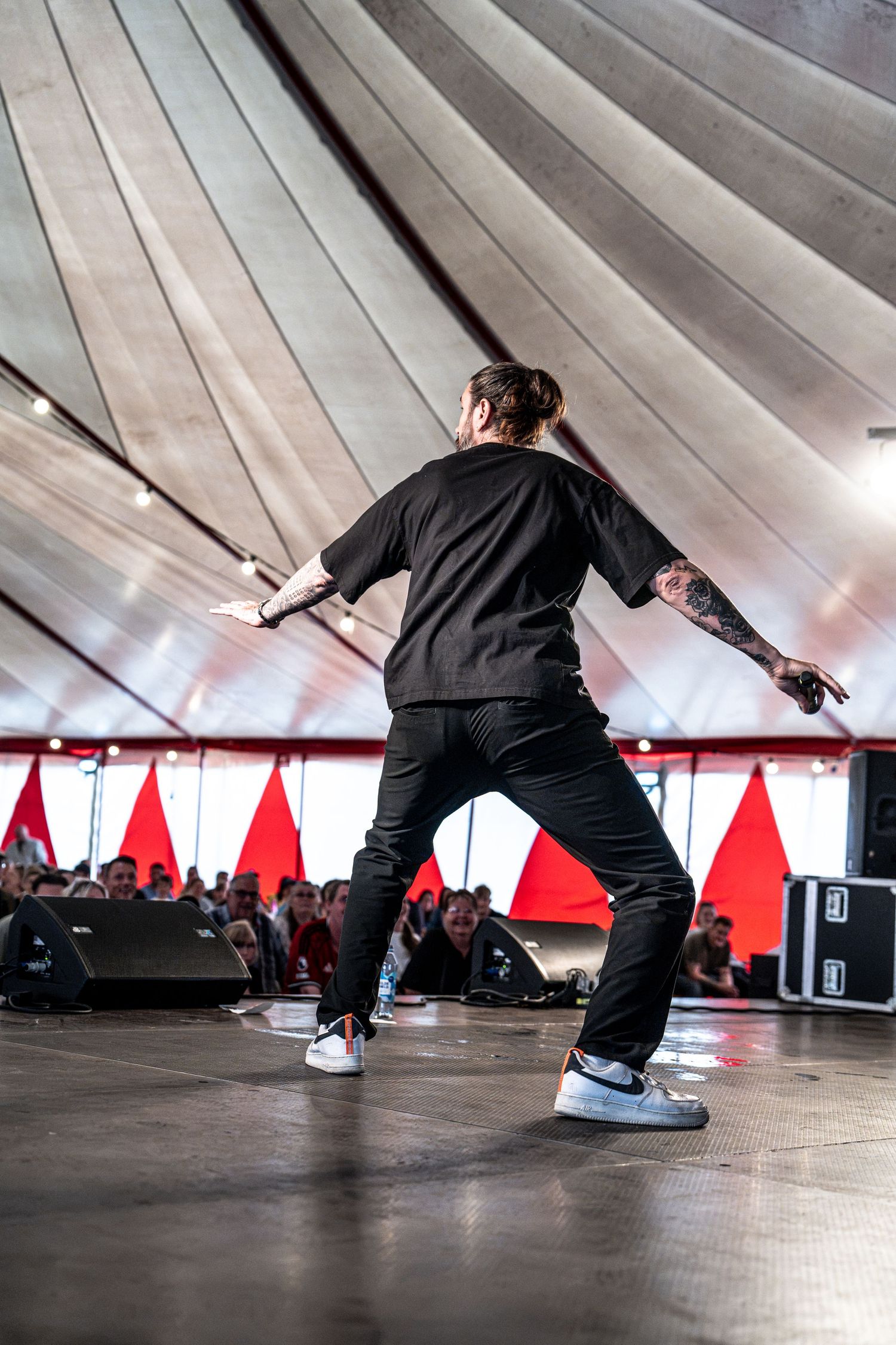 Tattooed rapper in black outfit performs energetic dance moves on stage at outdoor music festival.