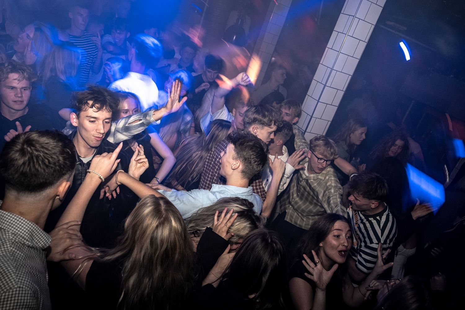 Energetic crowd of young partygoers dancing with raised hands in a dark nightclub lit by vivid blue and orange lights.