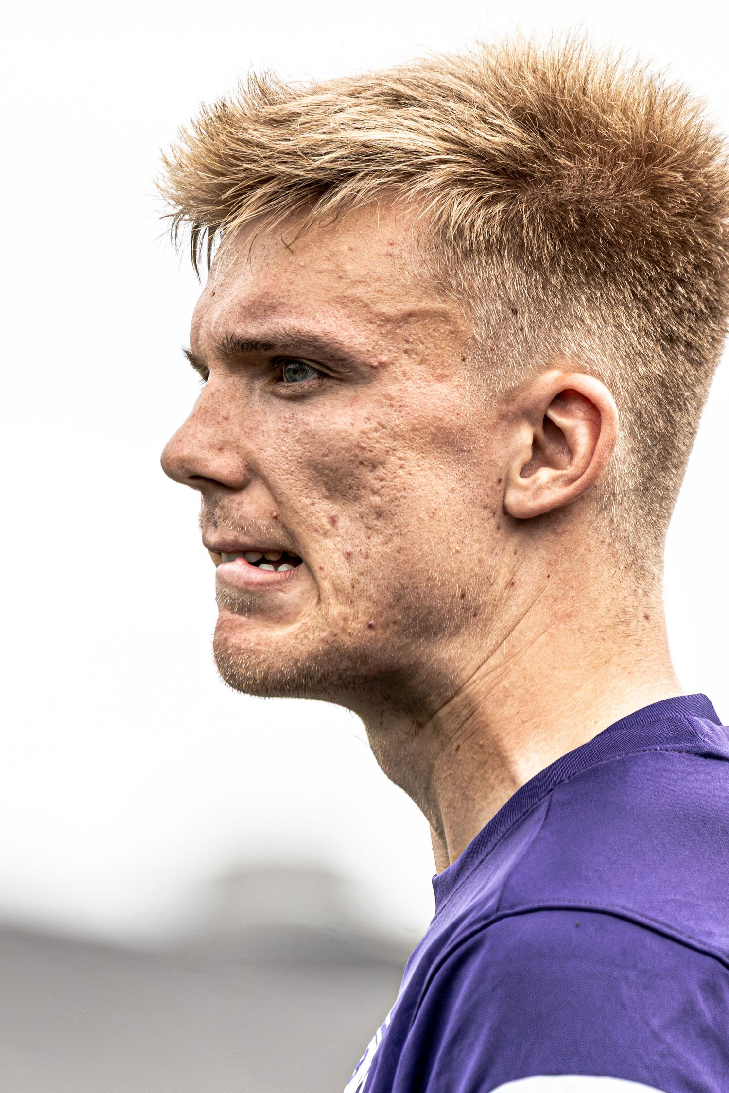 Close-up profile of a young blonde male athlete with freckles wearing a purple sports jersey outdoors.