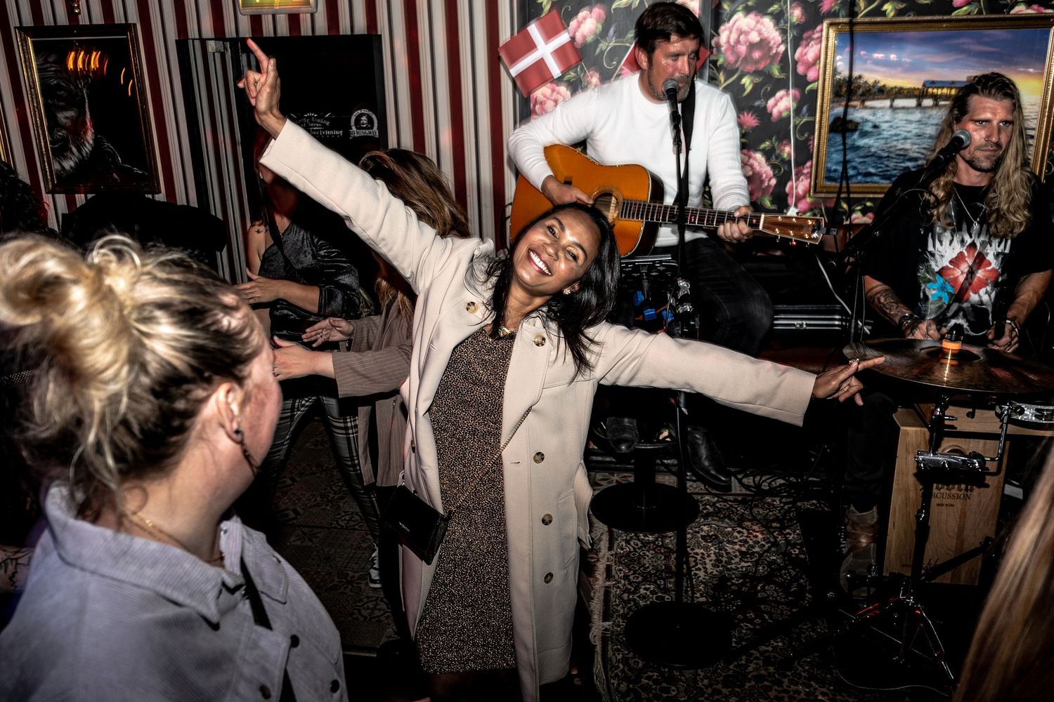 A joyful woman in a cream coat dances with arms outstretched as a guitarist and band perform at a crowded, colorful venue.