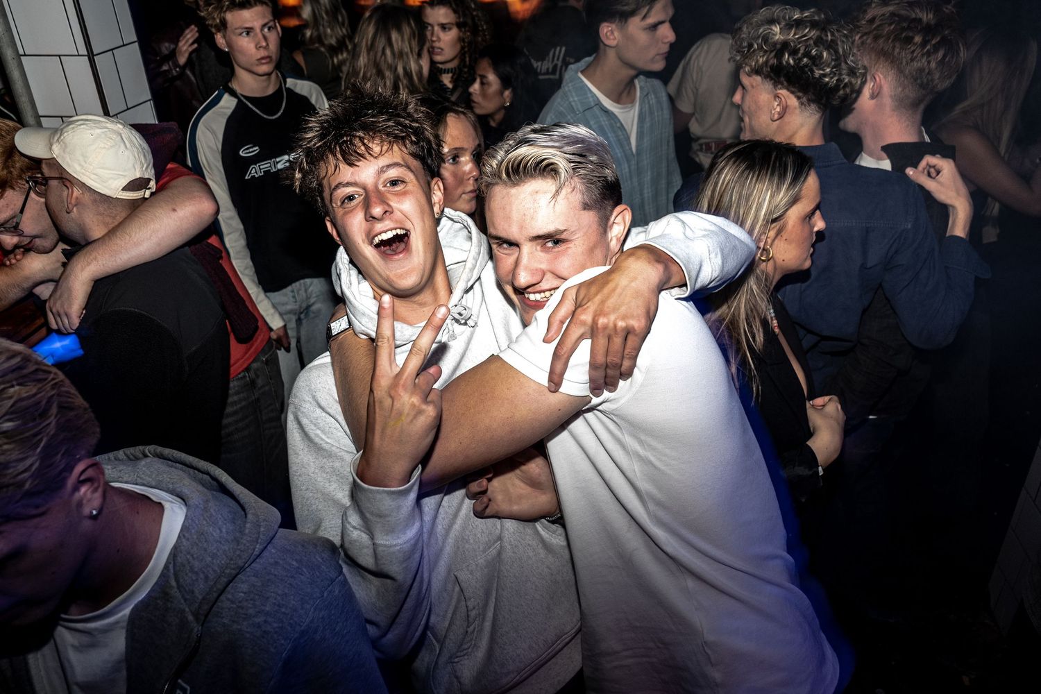 Two friends hug and laugh while posing for the camera at a crowded night party, one flashing a peace sign.