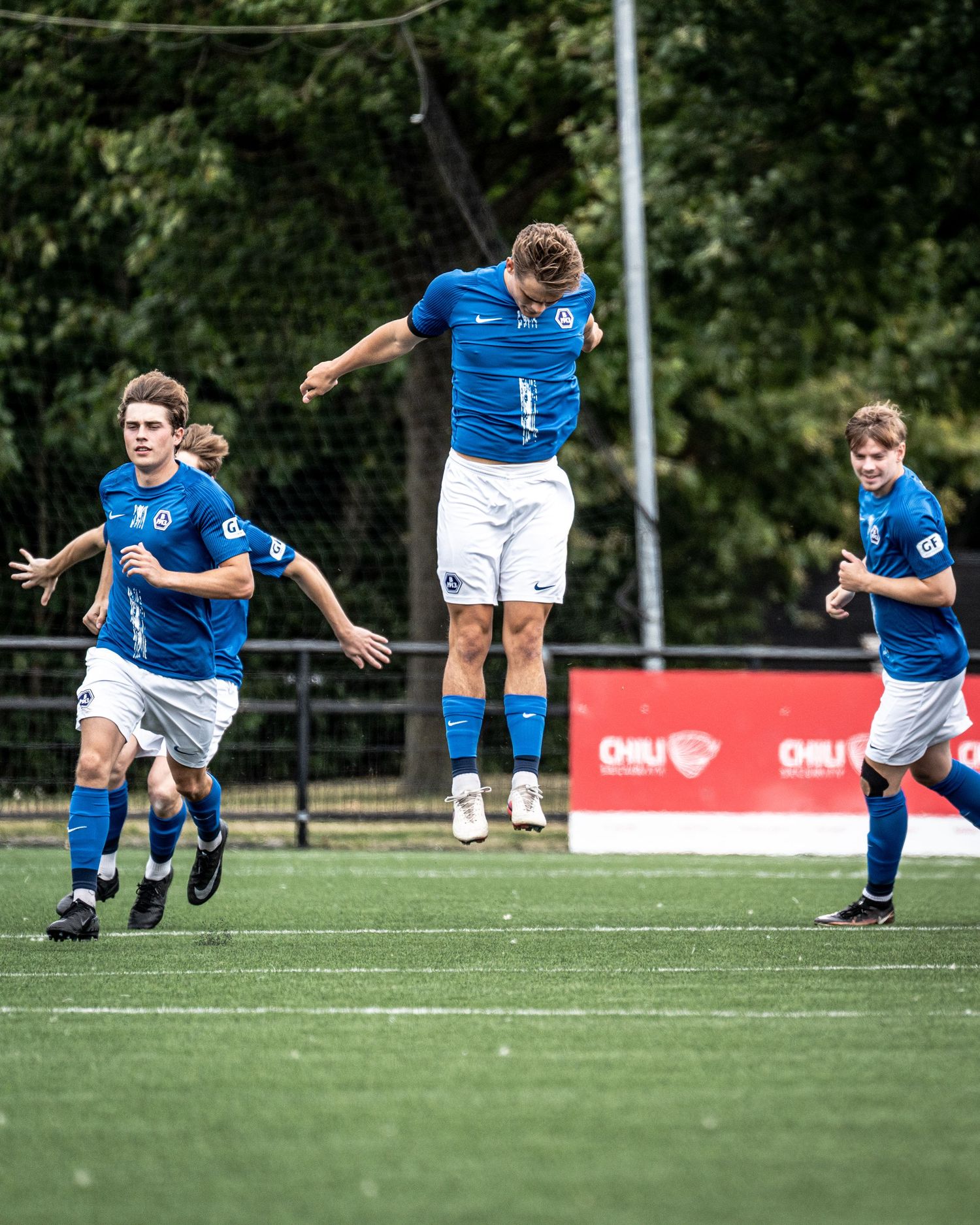 Soccer player jumps in celebration on green field while teammates in blue kits run toward him.