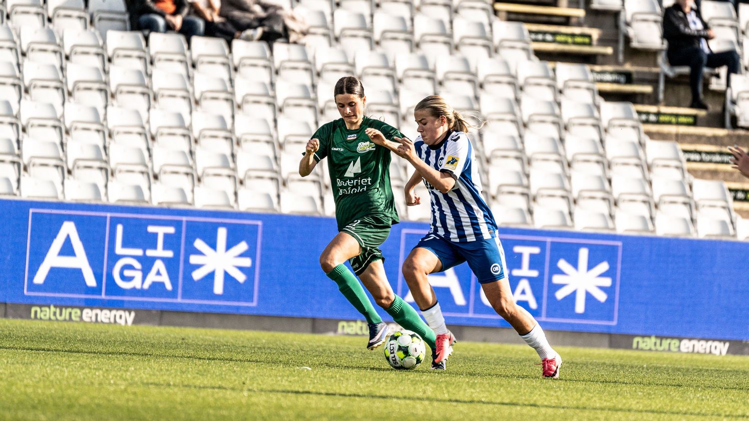 Two female footballers in green and blue and white kits battle for possession on stadium pitch.