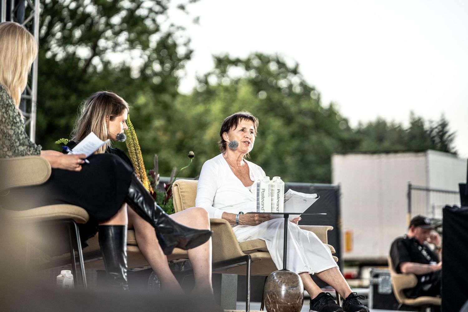 Three women engaged in a lively panel discussion on an outdoor stage surrounded by greenery.