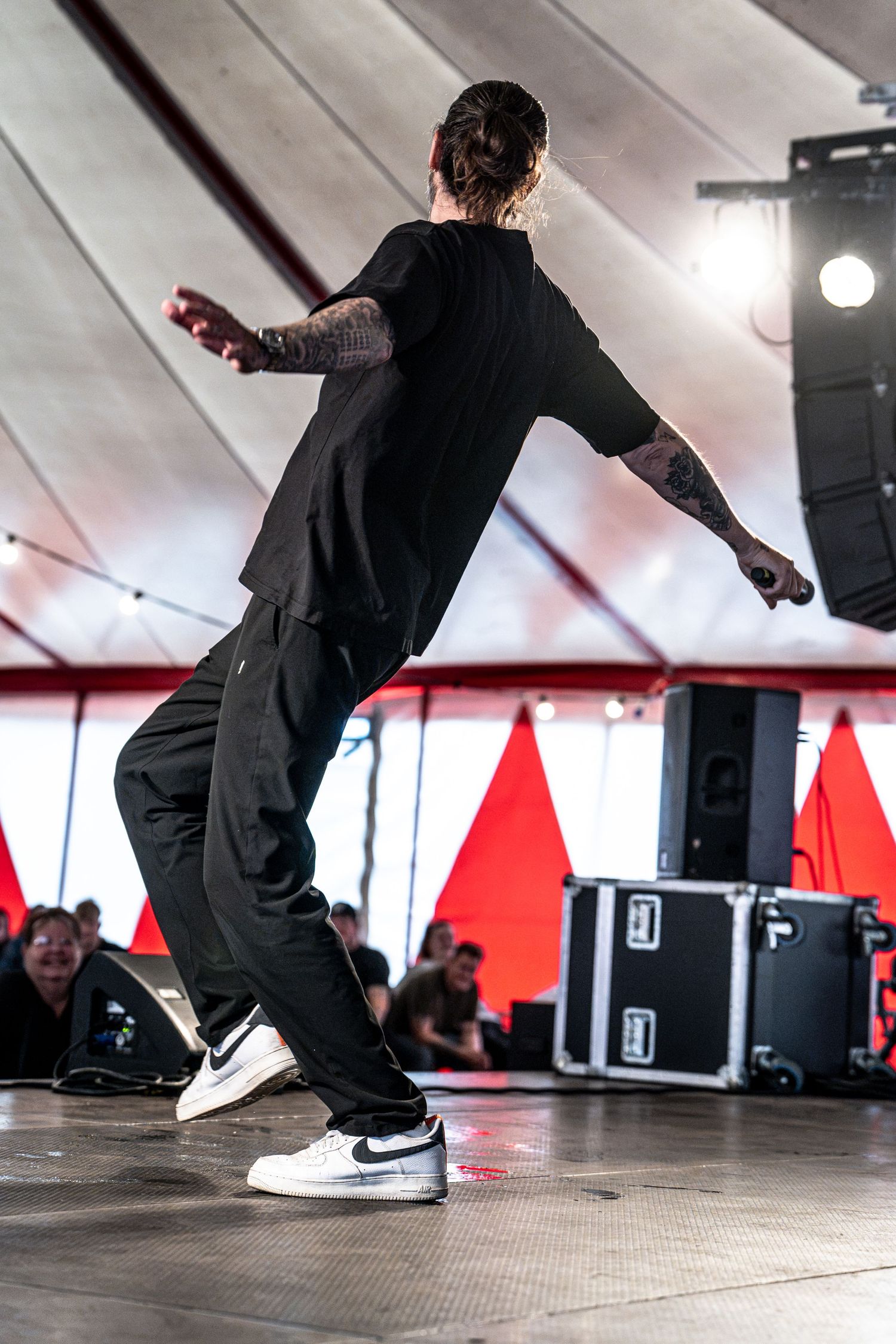 Tattooed performer in black outfit and Nike sneakers dances on festival stage from behind.