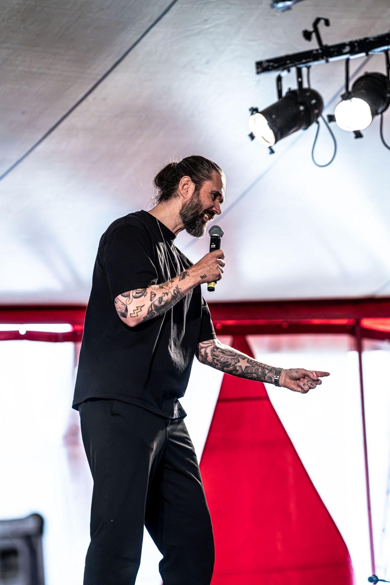 Tattooed male singer points to crowd while performing on stage with red backdrop.