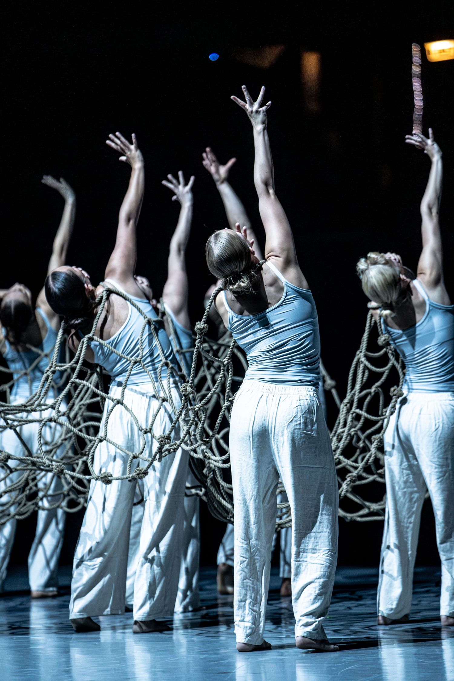 Group of dancers in white costumes holding rope nets with arms raised during a theatrical stage performance.