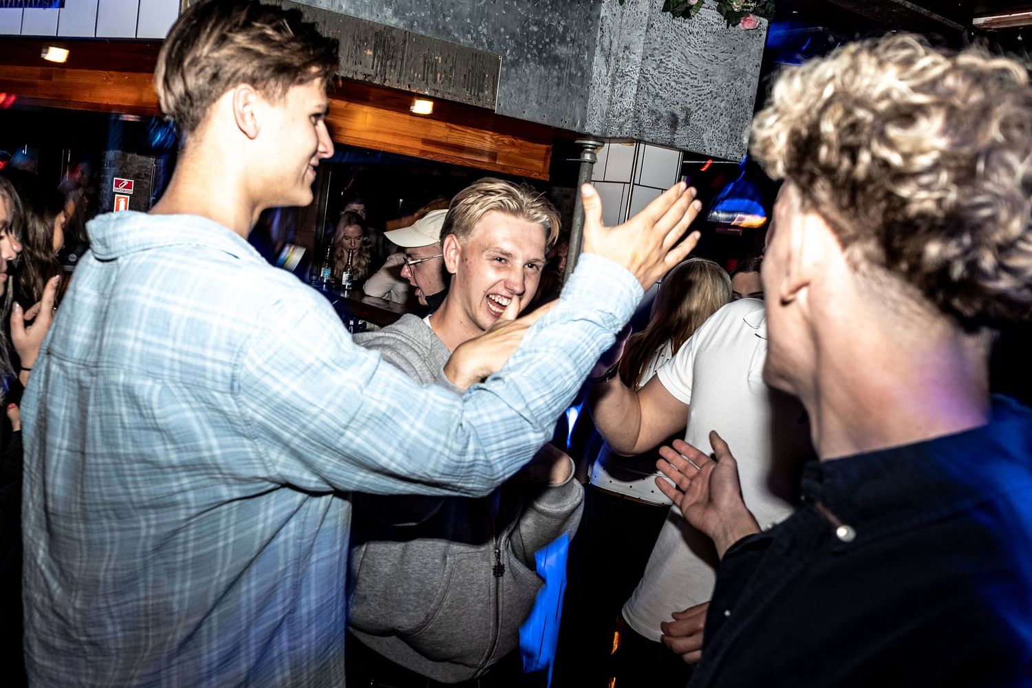 Young men laugh and interact on a crowded dance floor at a nightlife venue, with warm lights and partygoers in the background.