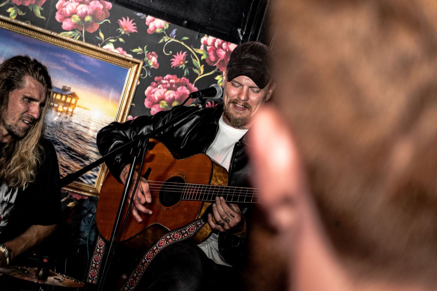 A bearded musician in a cap plays an acoustic guitar and sings into a microphone beside a long-haired man in a cozy venue.