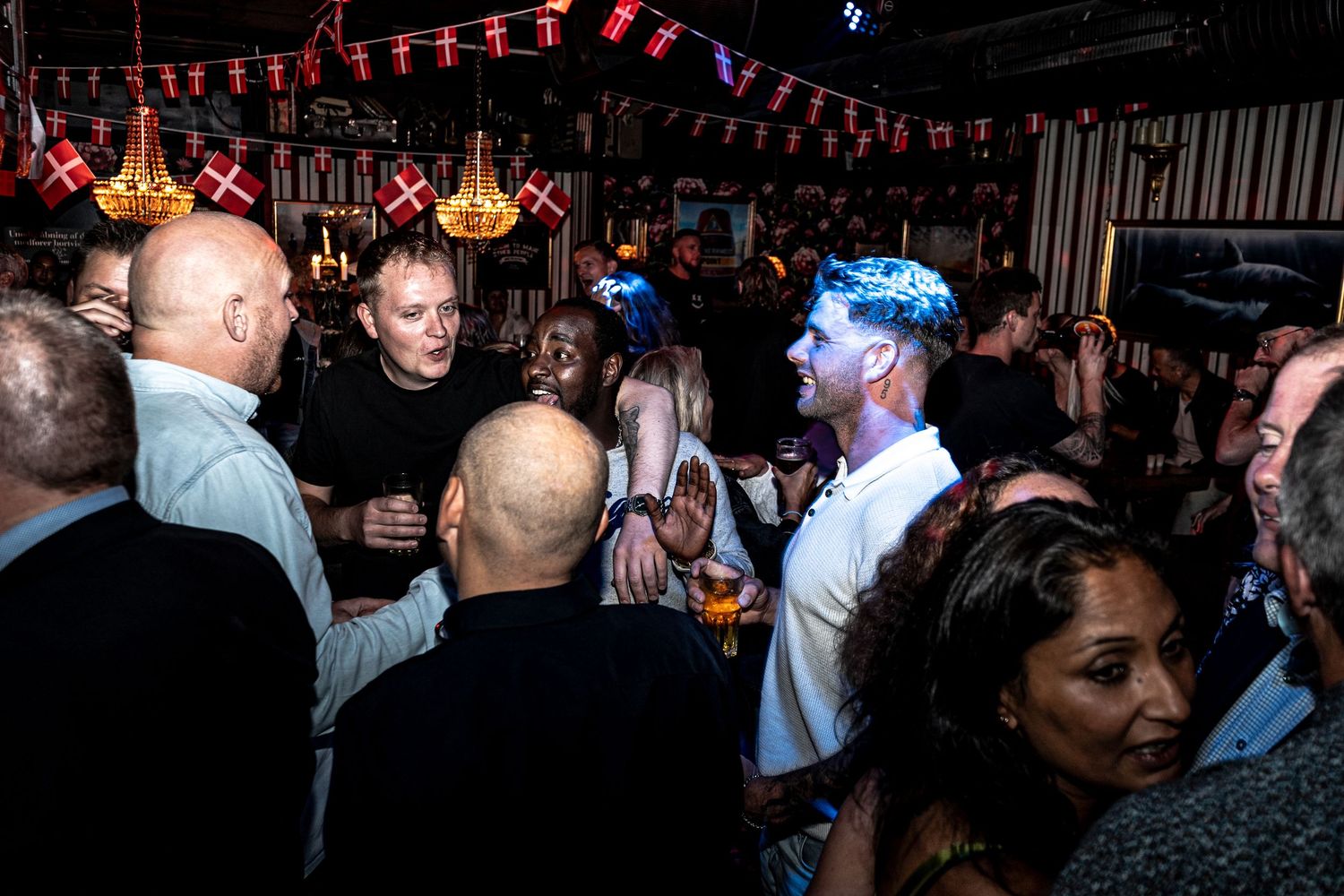 Lively crowd of people chatting and drinking at a festive bar decorated with hanging lanterns and small flags.