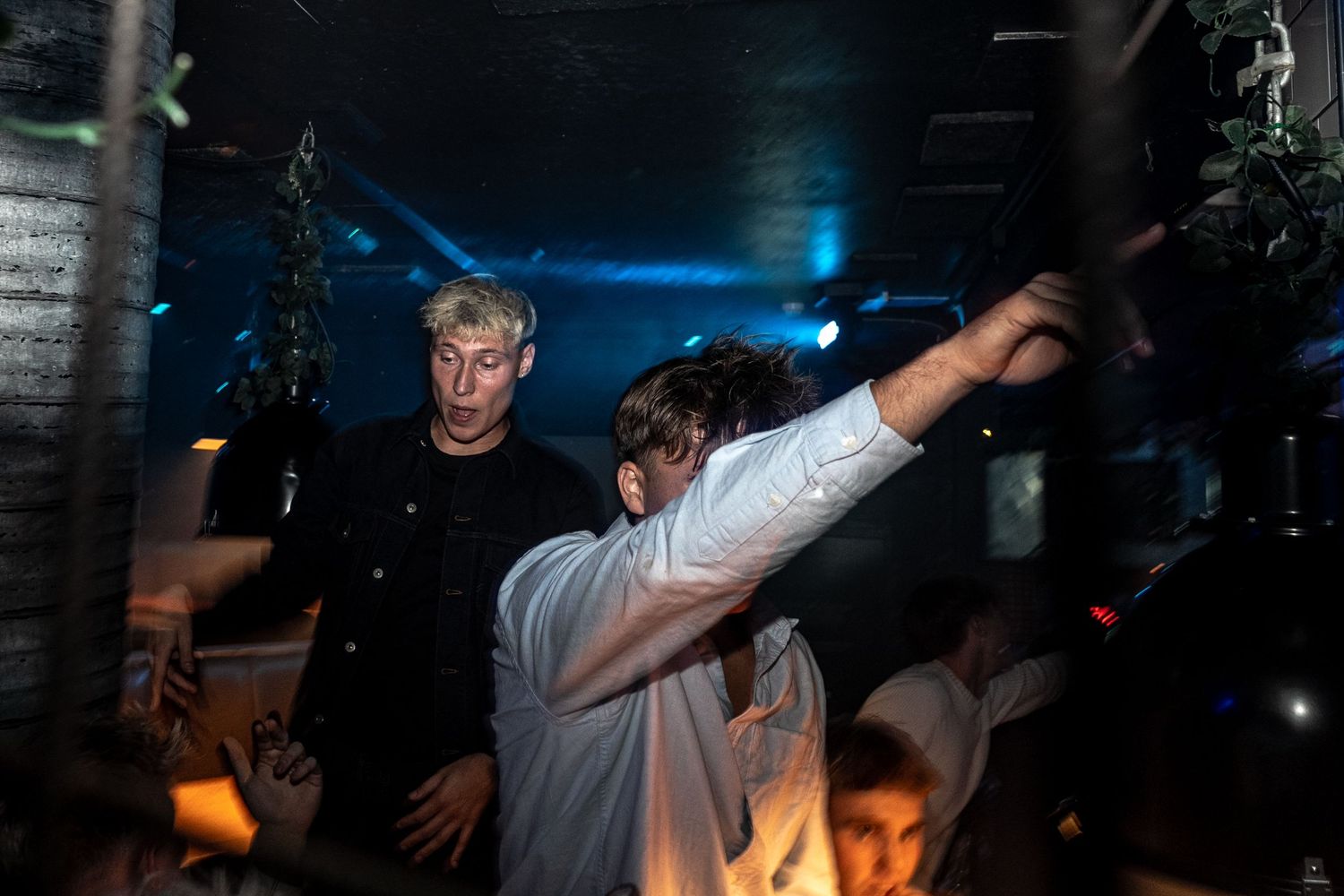 Partygoers dance in a dark, moody nightclub as a man in a white shirt raises his arm while another looks on with surprise.