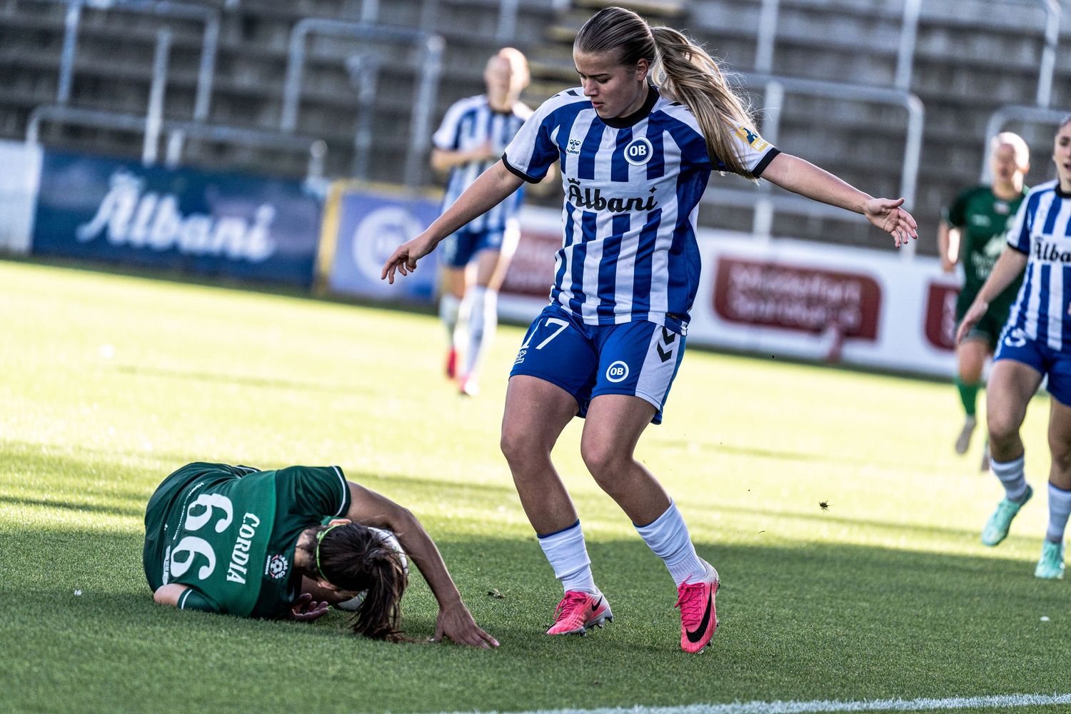 Female soccer player in blue and white kit stepping over fallen opponent on field.