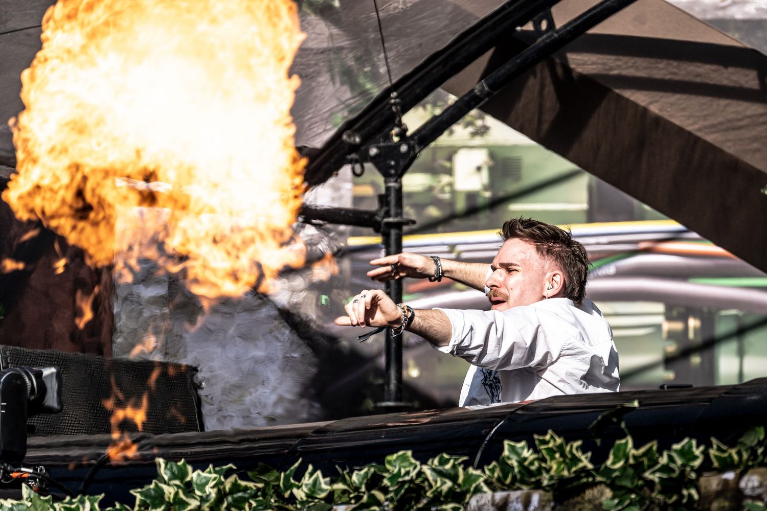 DJ performing at outdoor stage with dramatic fire pyrotechnics explosion behind equipment.