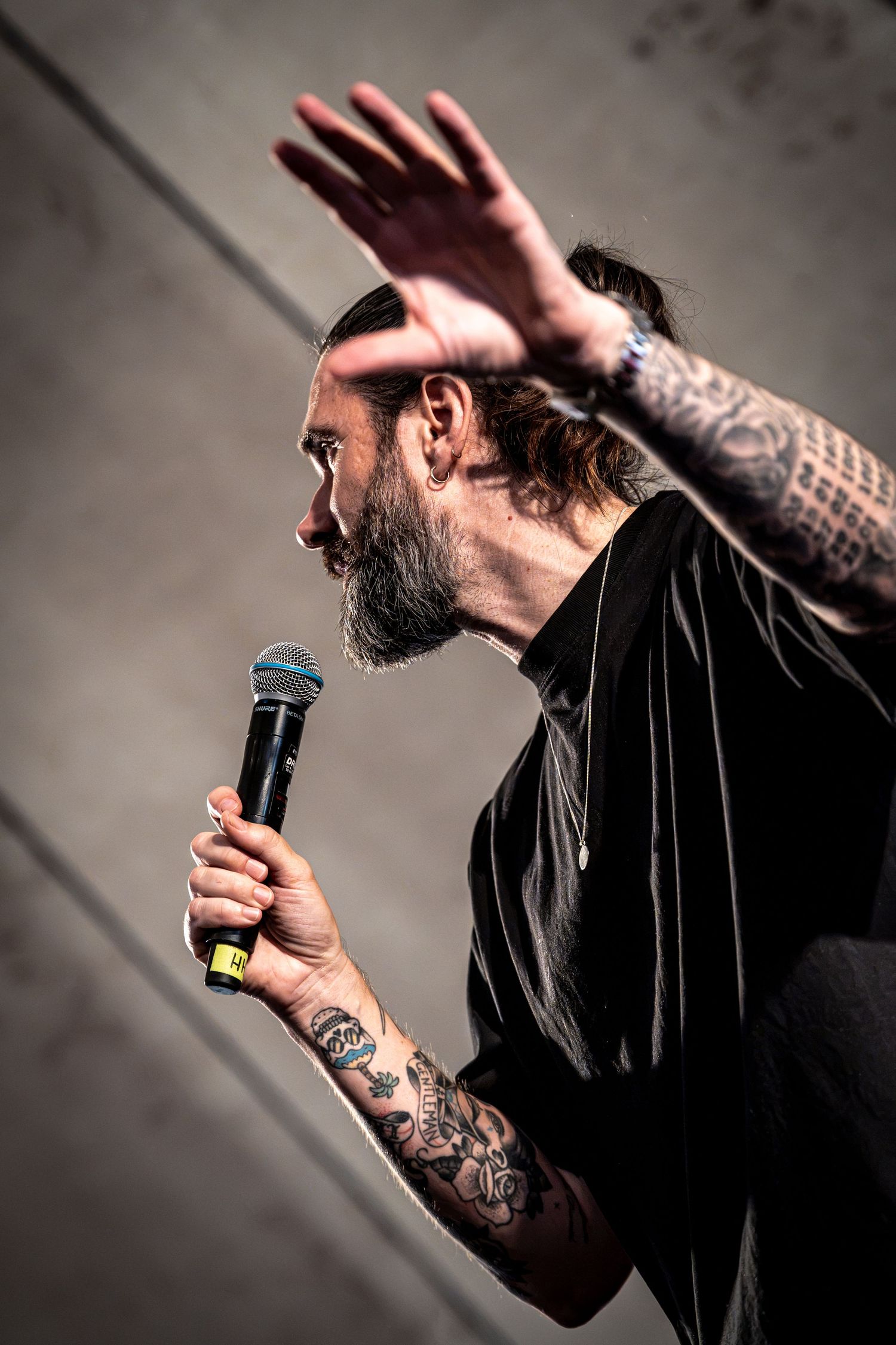 Tattooed bearded performer raising hand while holding microphone in profile view on stage.