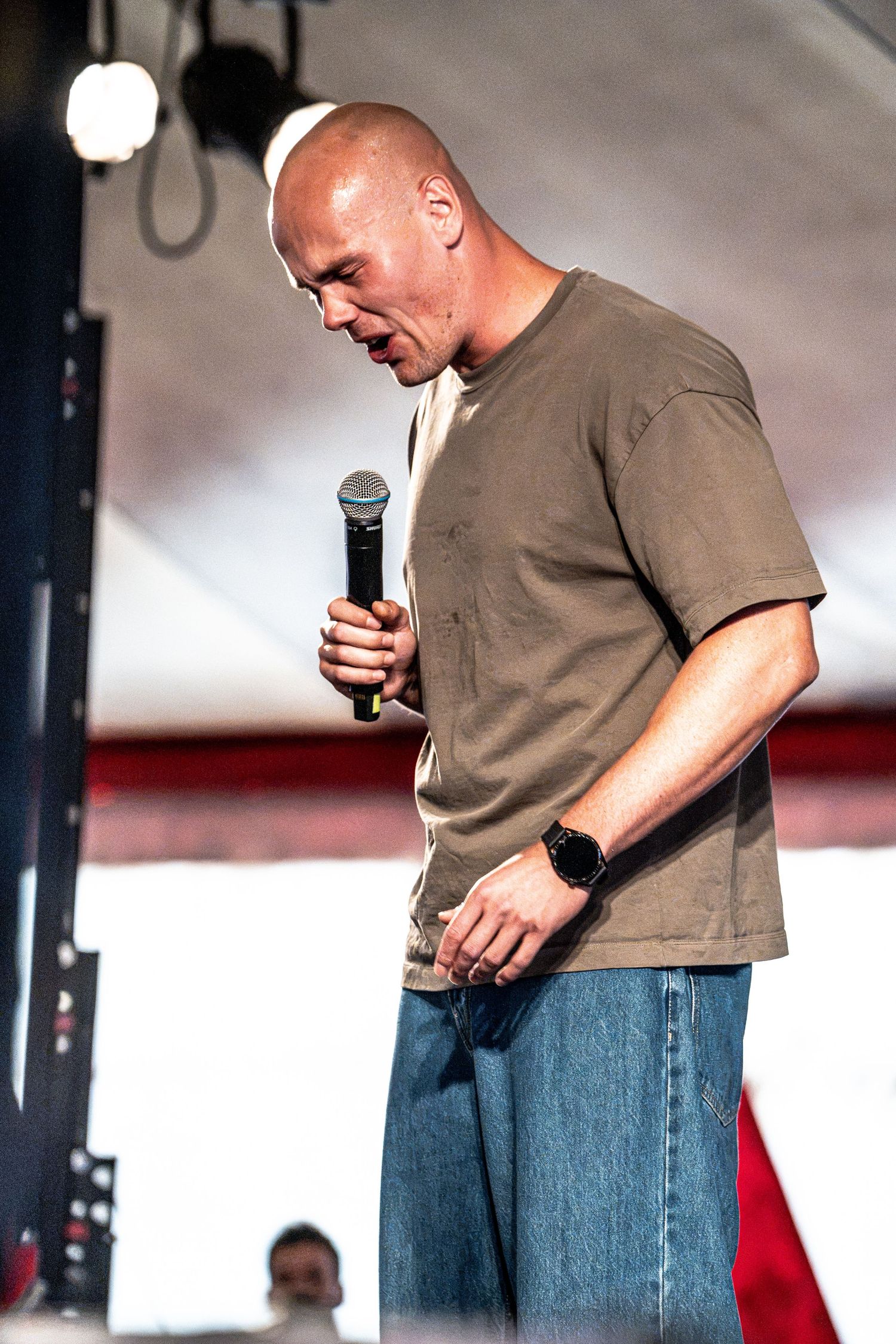 Bald male speaker holds microphone on stage, head bowed, wearing olive t-shirt and jeans at outdoor festival.